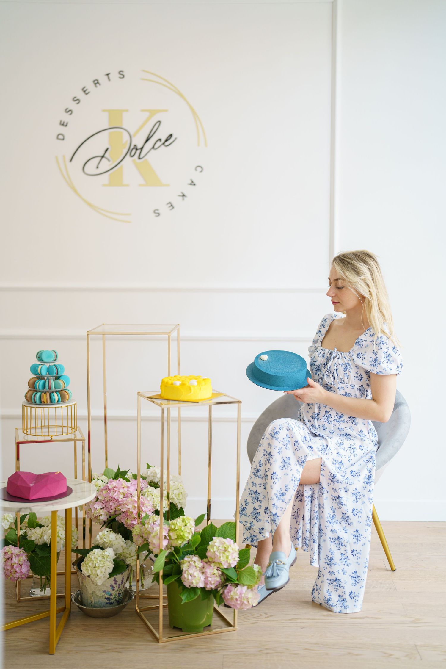 Blonde woman in floral dress holding blue cake at Dolce K Desserts bakery with colorful cakes and hydrangeas.