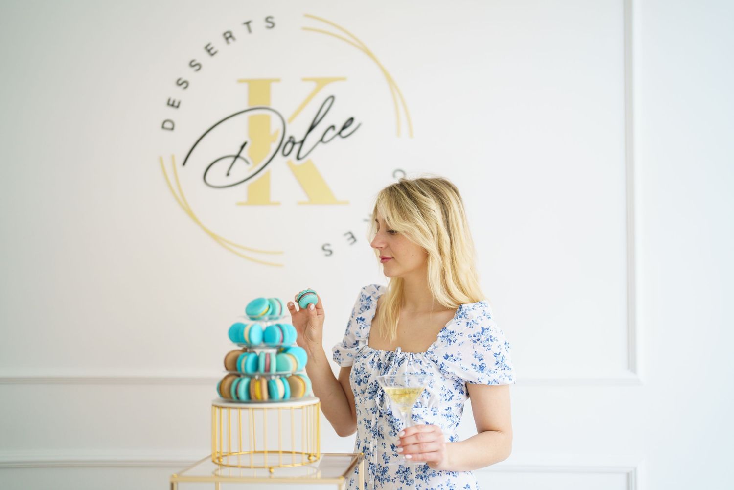 Woman in floral dress holding macaron beside tiered tower at Dolce K Desserts Studio event.