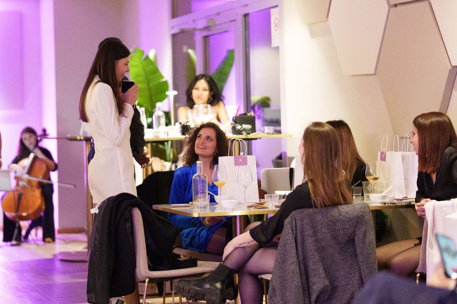 Women socializing at an elegant purple-lit event venue with live cello music in the background.