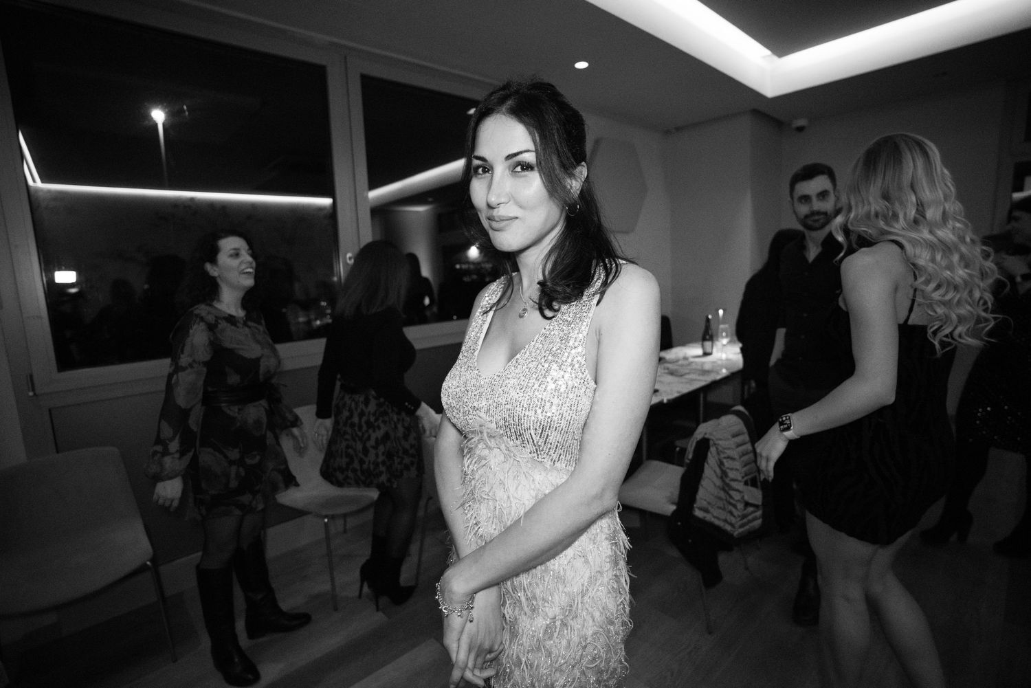 Black and white photo of an elegant woman in a sequined dress at a social gathering with guests mingling in the background.