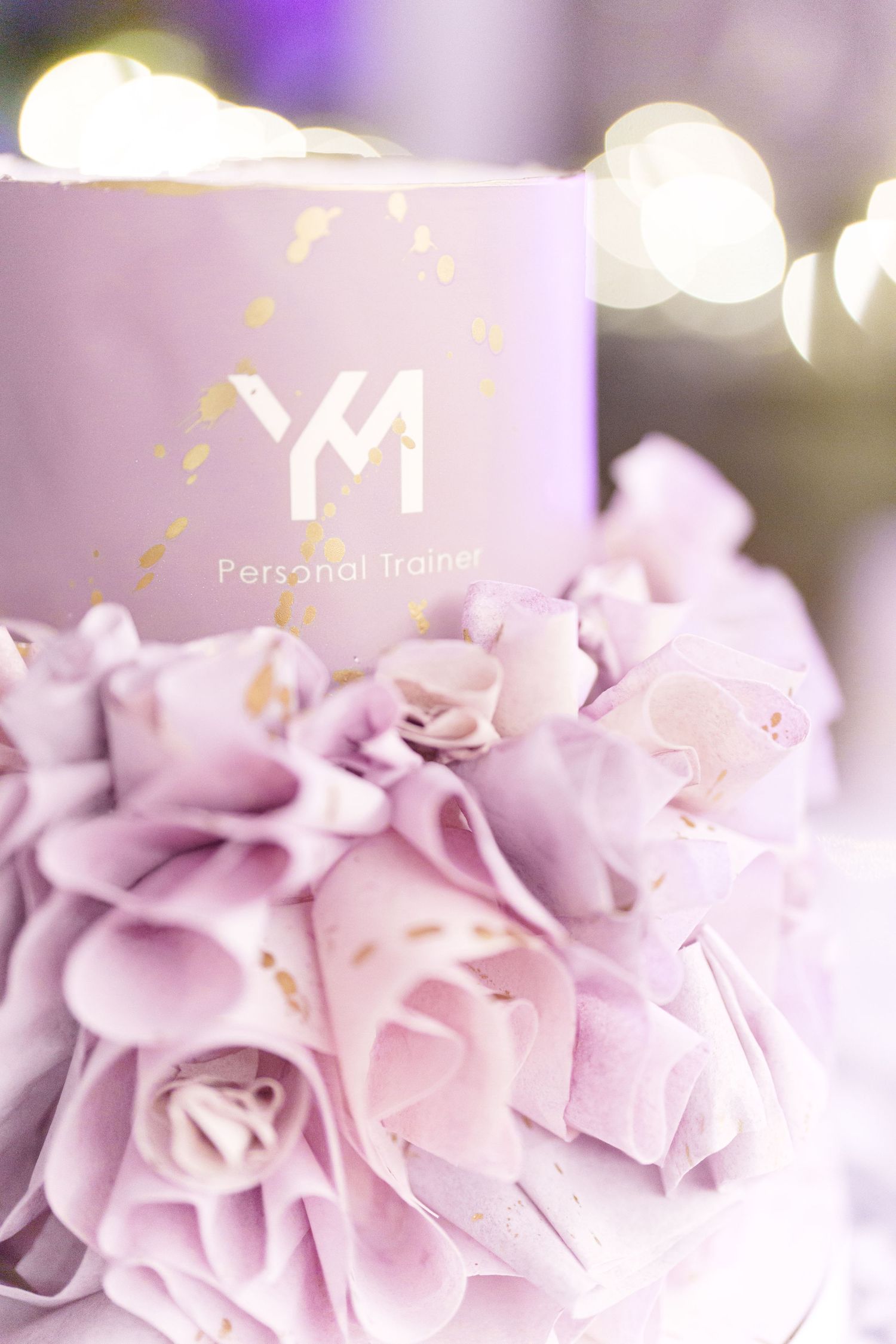 Elegant lavender tiered cake with gold YM Personal Trainer logo and ruffled fondant flower decorations.