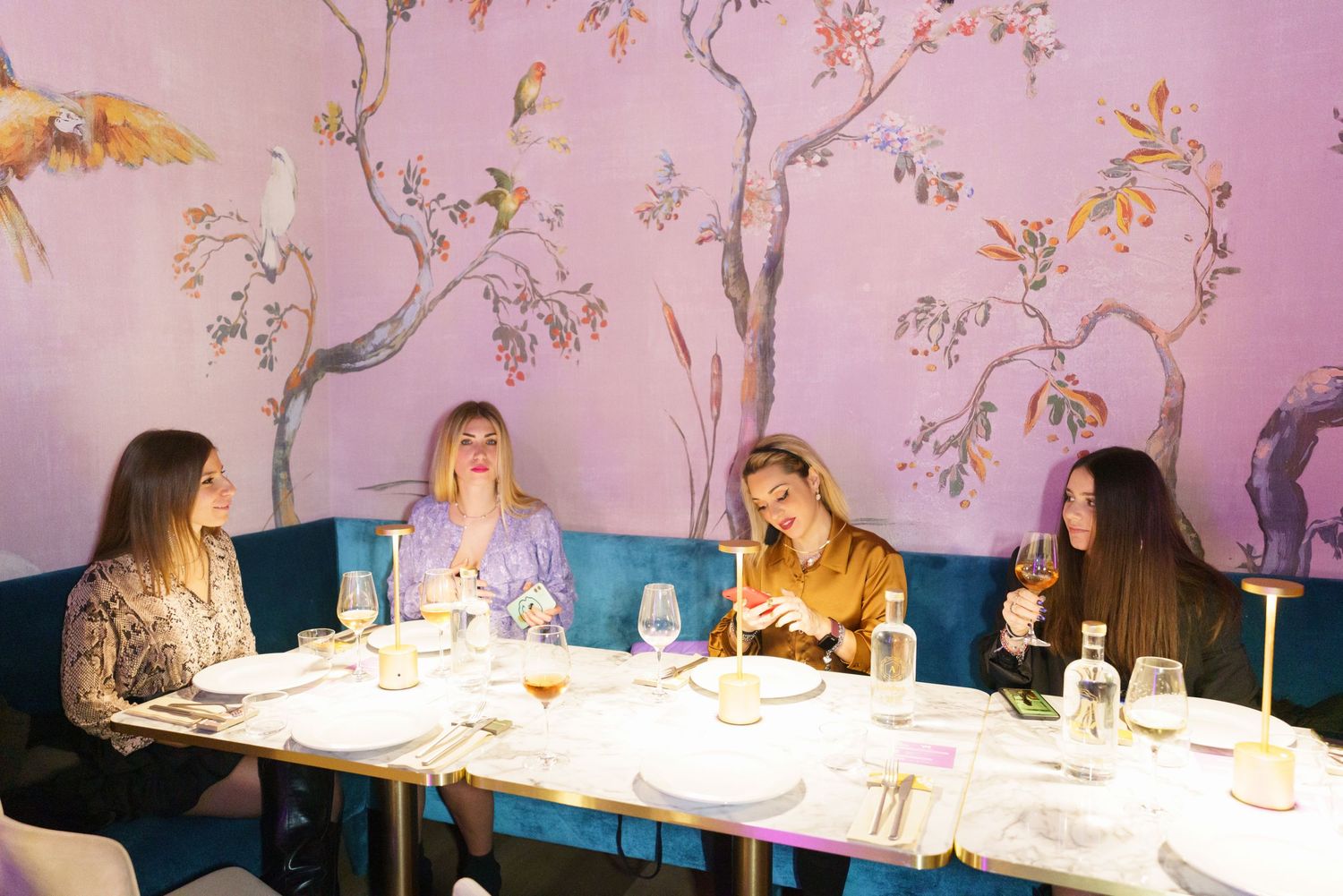 Four women dining at an elegant restaurant with floral mural walls, fine china, and wine glasses on a white tablecloth.
