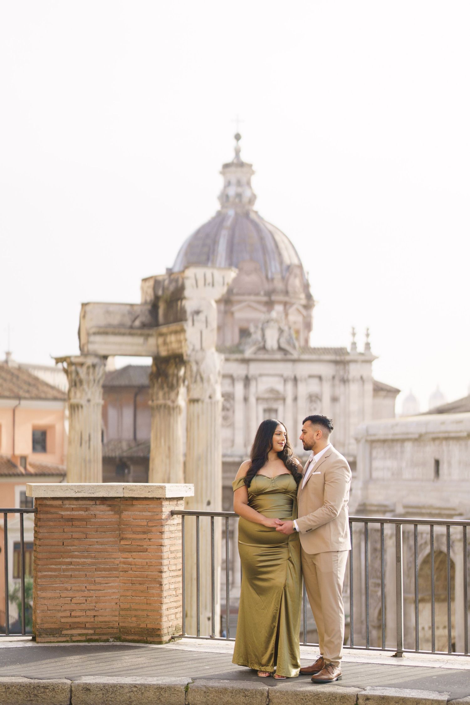 Couple in elegant attire embracing on a Roman terrace with ancient columns and a domed church backdrop.