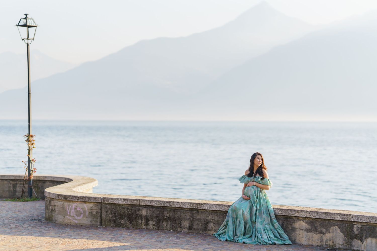 Woman in teal gown sits on lakeside wall with misty mountains and calm water in background.