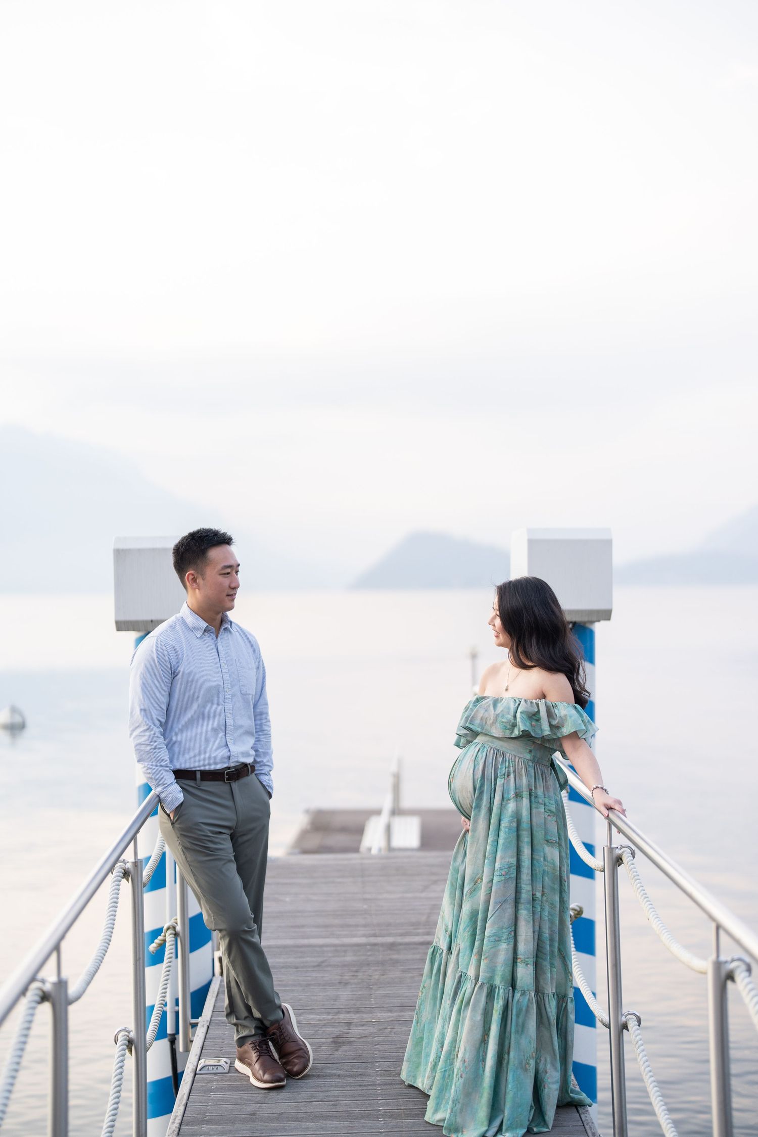 Pregnant couple standing on a waterfront pier with mountain views, woman in teal off-shoulder gown, man in light blue shirt.