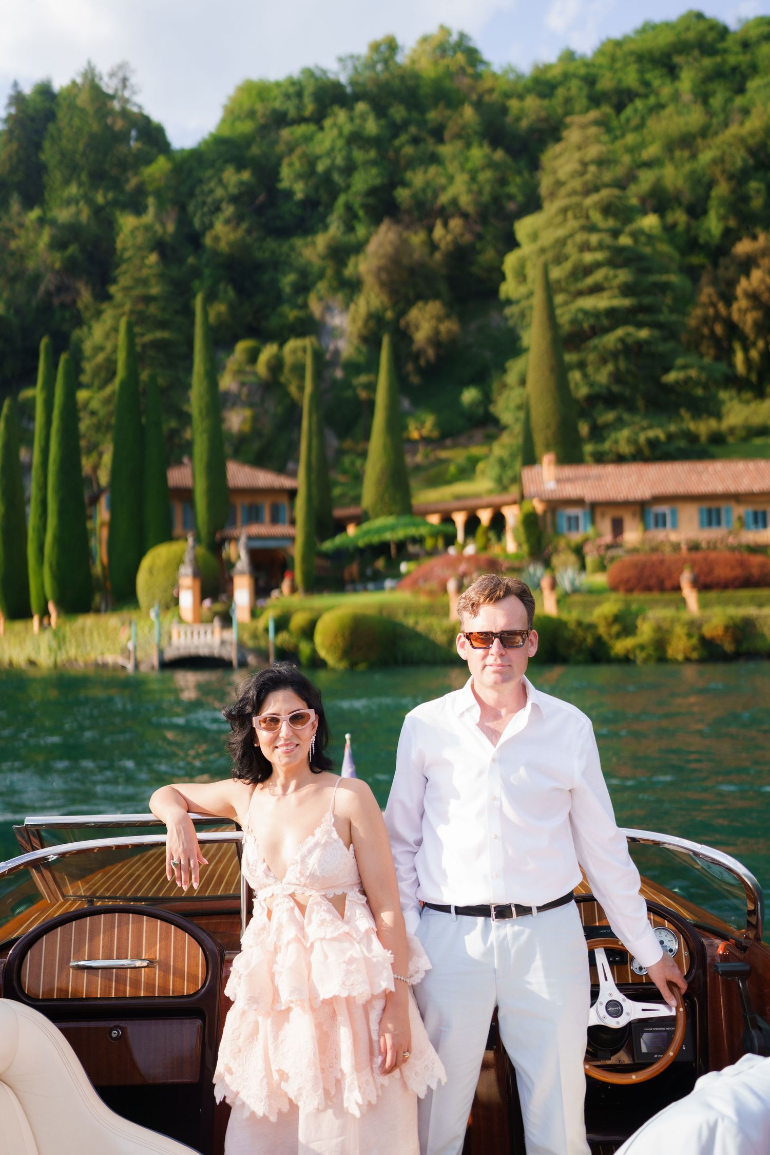 Couple in elegant attire on a wooden boat with lush Italian villa and cypress trees on Lake Como.