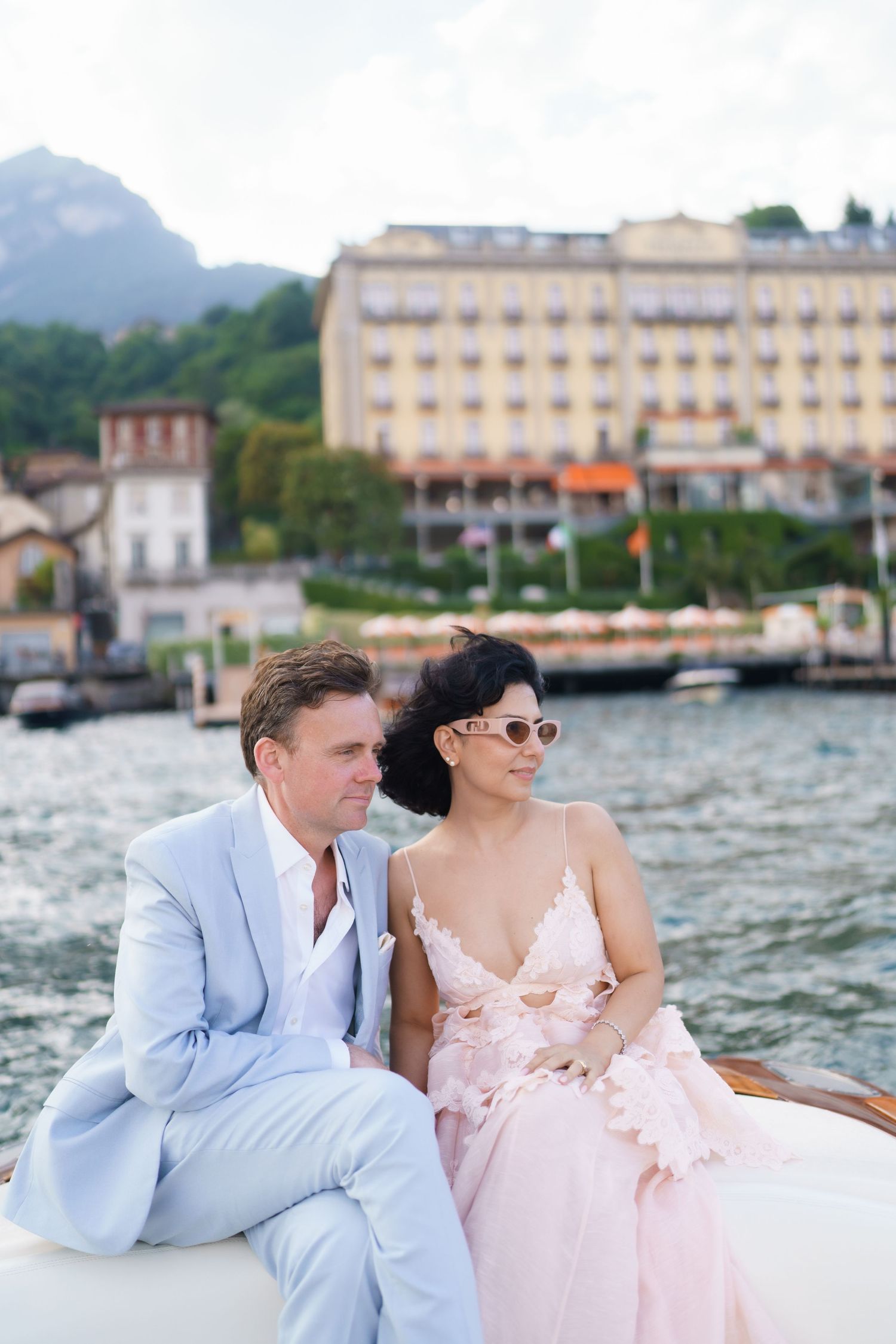 Elegant couple in formal attire sitting on a boat with a grand lakeside hotel and mountains in the background.
