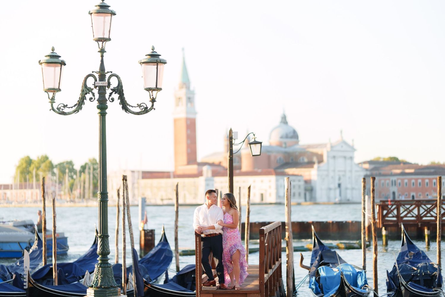 Couple posing by gondolas on Venice waterfront with San Giorgio Maggiore church in background at sunset.