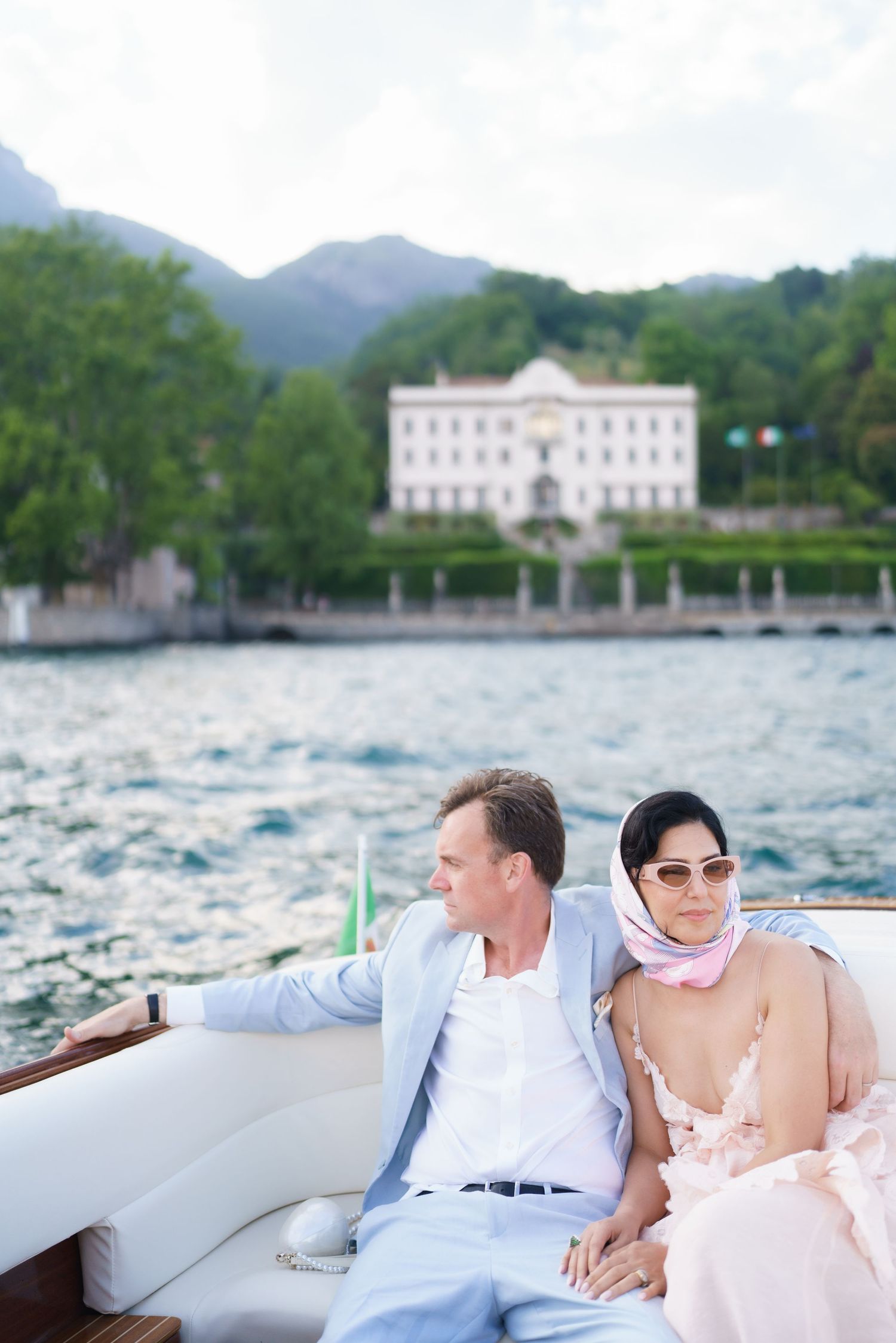 Elegant couple relaxing on a luxury boat on Lake Como, Italy, with a grand villa and mountains in the background.