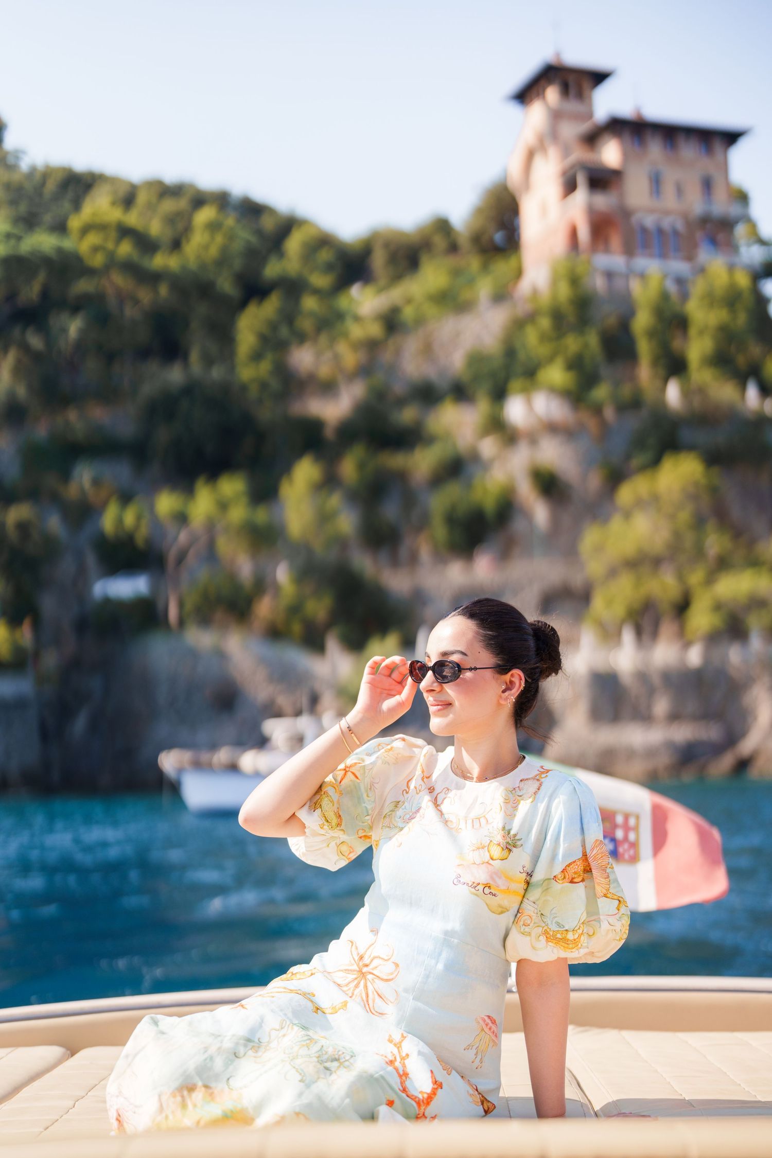Elegant woman in floral dress and sunglasses on a yacht with a Mediterranean villa and lush hillside backdrop.
