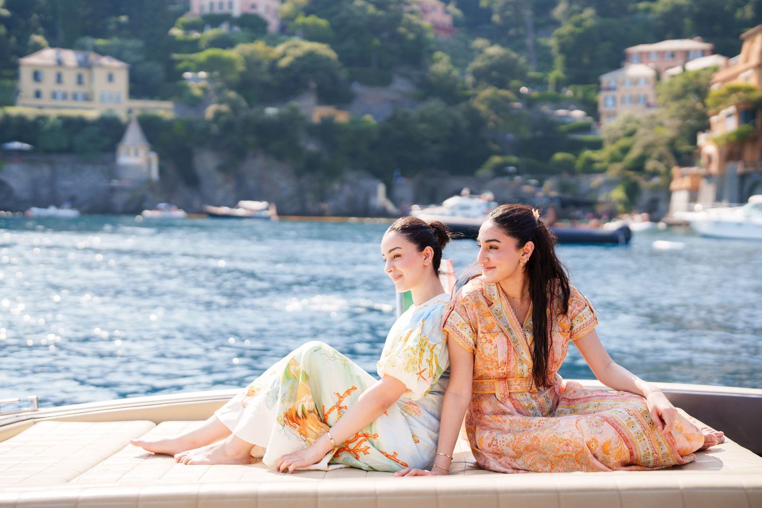 Two women in floral dresses sit back-to-back on a boat deck with a scenic coastal village and blue water behind them.