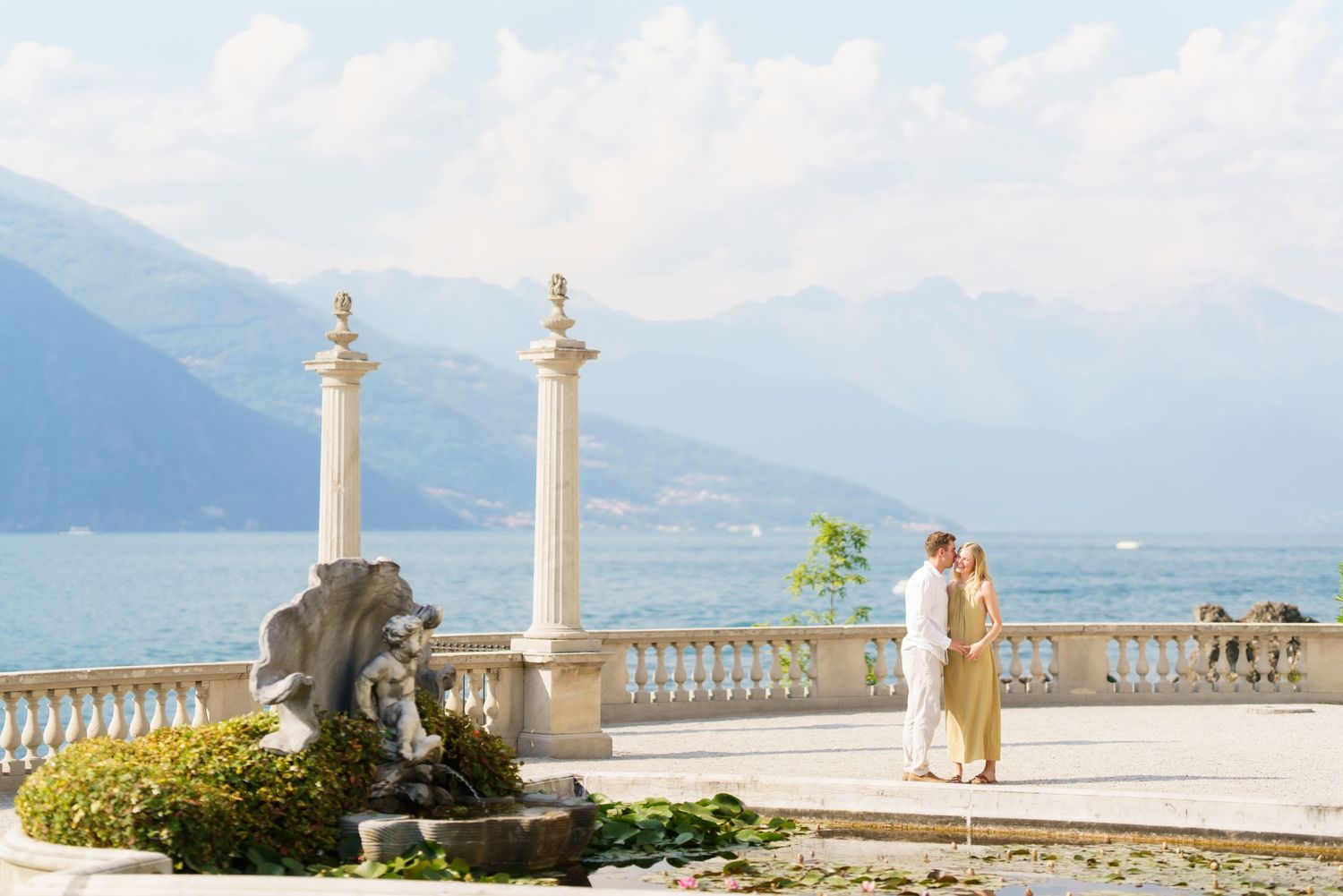 Couple embracing on elegant lakeside terrace with marble columns, fountain, and stunning mountain backdrop at Lake Como.
