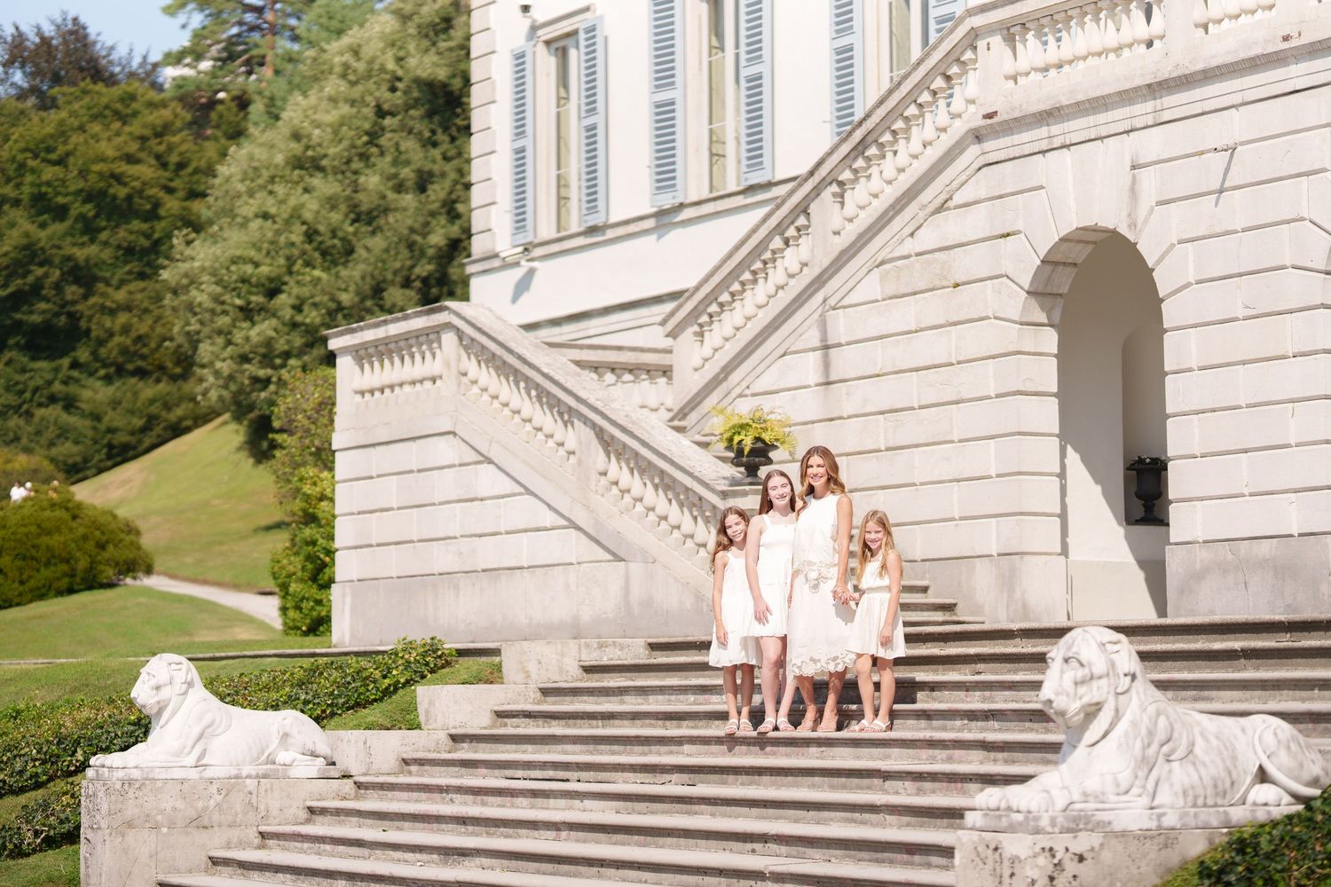 Group of women and girls in white dresses posing on grand stone staircase of elegant villa with lion sculptures.