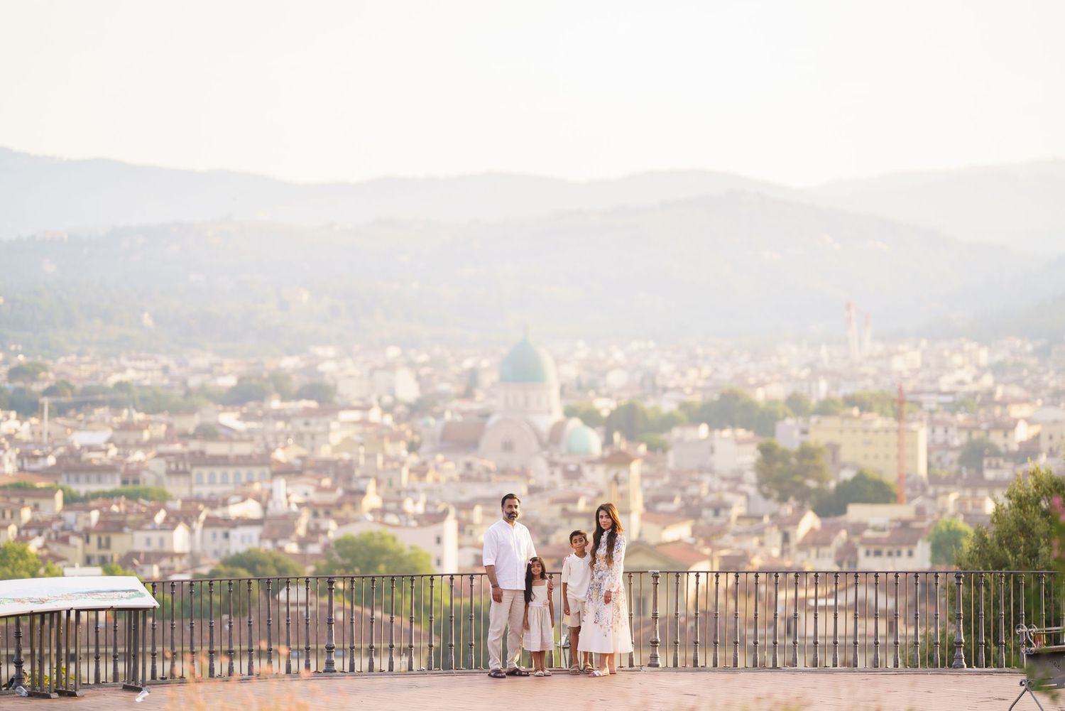 Family in white outfits posing at Florence overlook with panoramic city view and mountains in background.