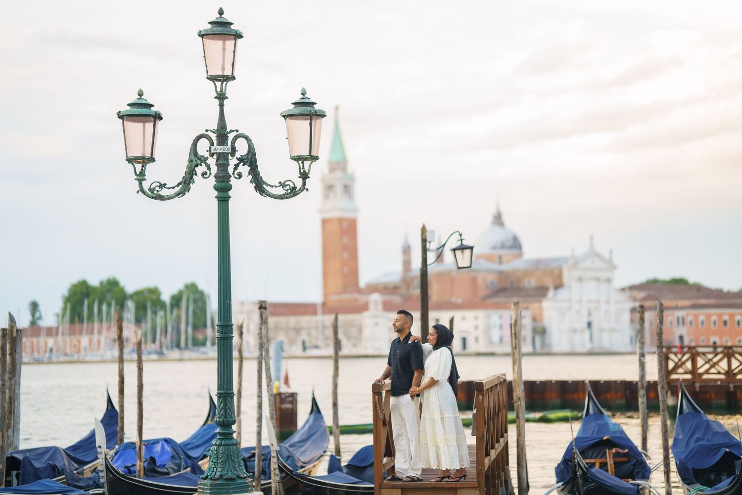 Couple standing near gondolas on Venice waterfront with San Giorgio Maggiore church in background at sunset.