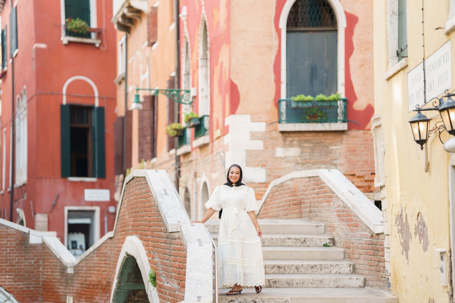 Woman in white dress standing on brick bridge steps in Venice, Italy, with colorful buildings backdrop.