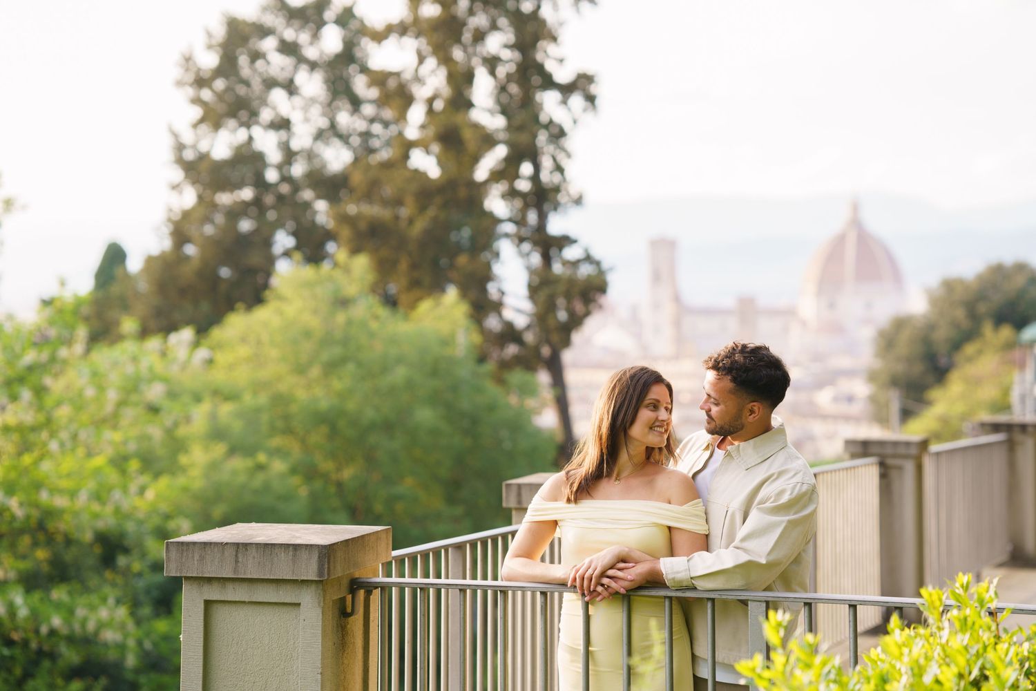 Couple embracing on a balcony with Florence cathedral dome and lush green trees in the background.