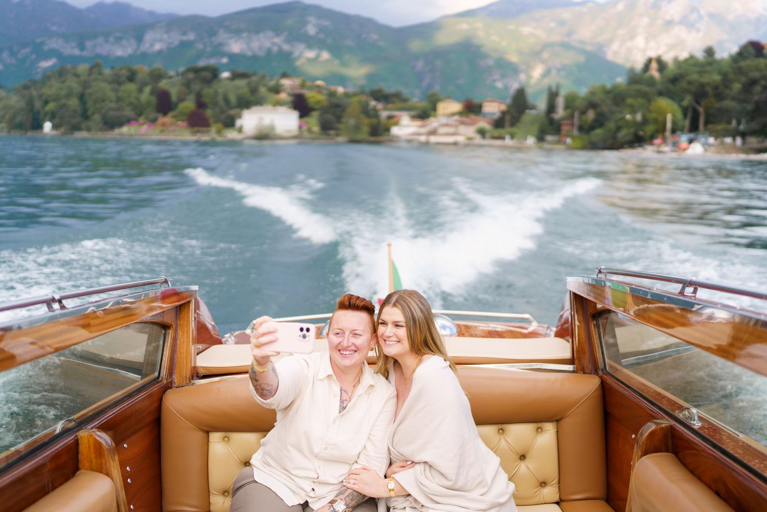 Two women taking a selfie on a luxury wooden speedboat on a scenic mountain lake.