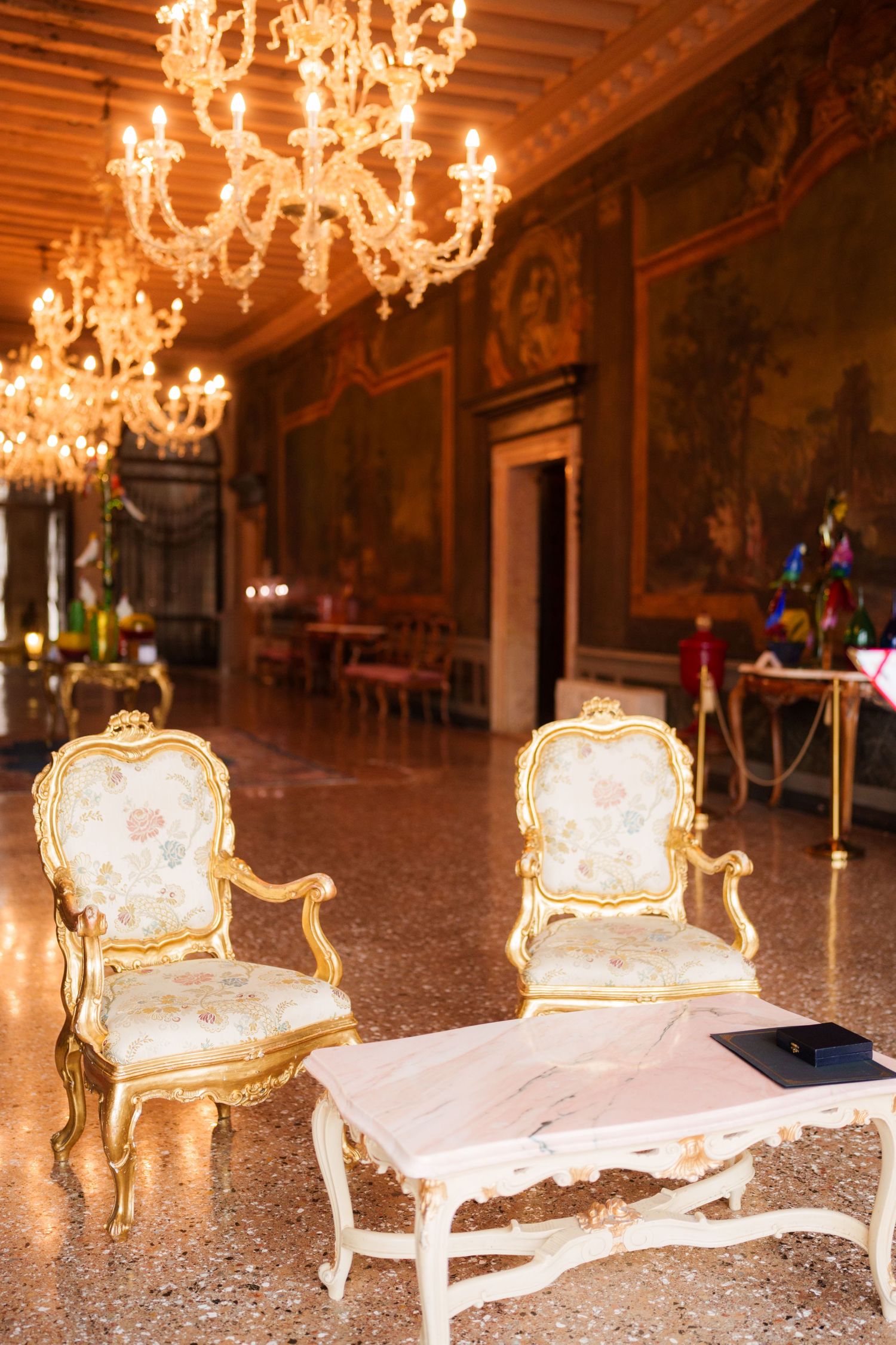 Ornate antique chairs and benches arranged in a luxurious room with crystal chandeliers and decorative wall murals.