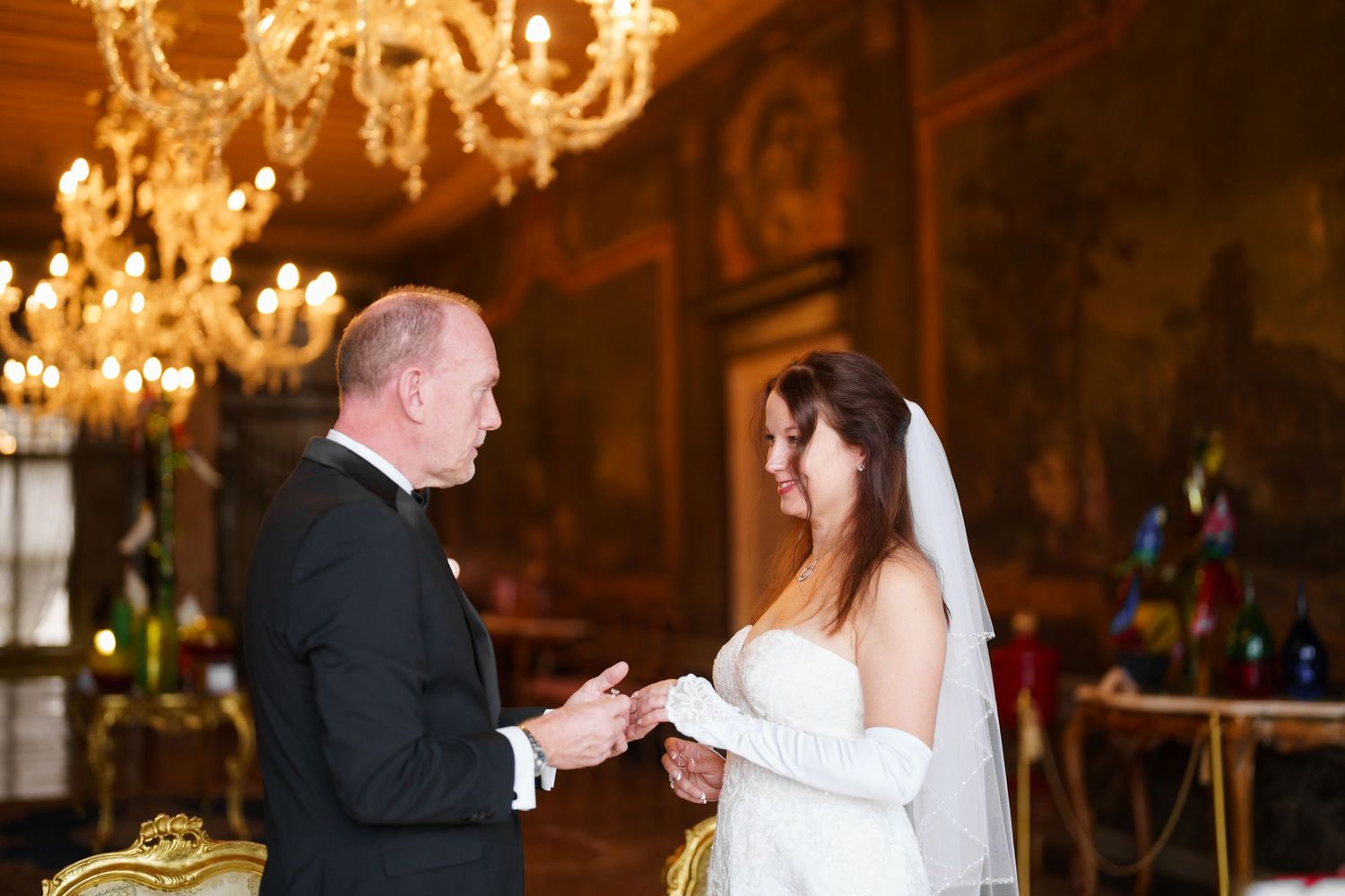 A wedding ceremony takes place in an elegant ballroom with ornate chandeliers and warm lighting.