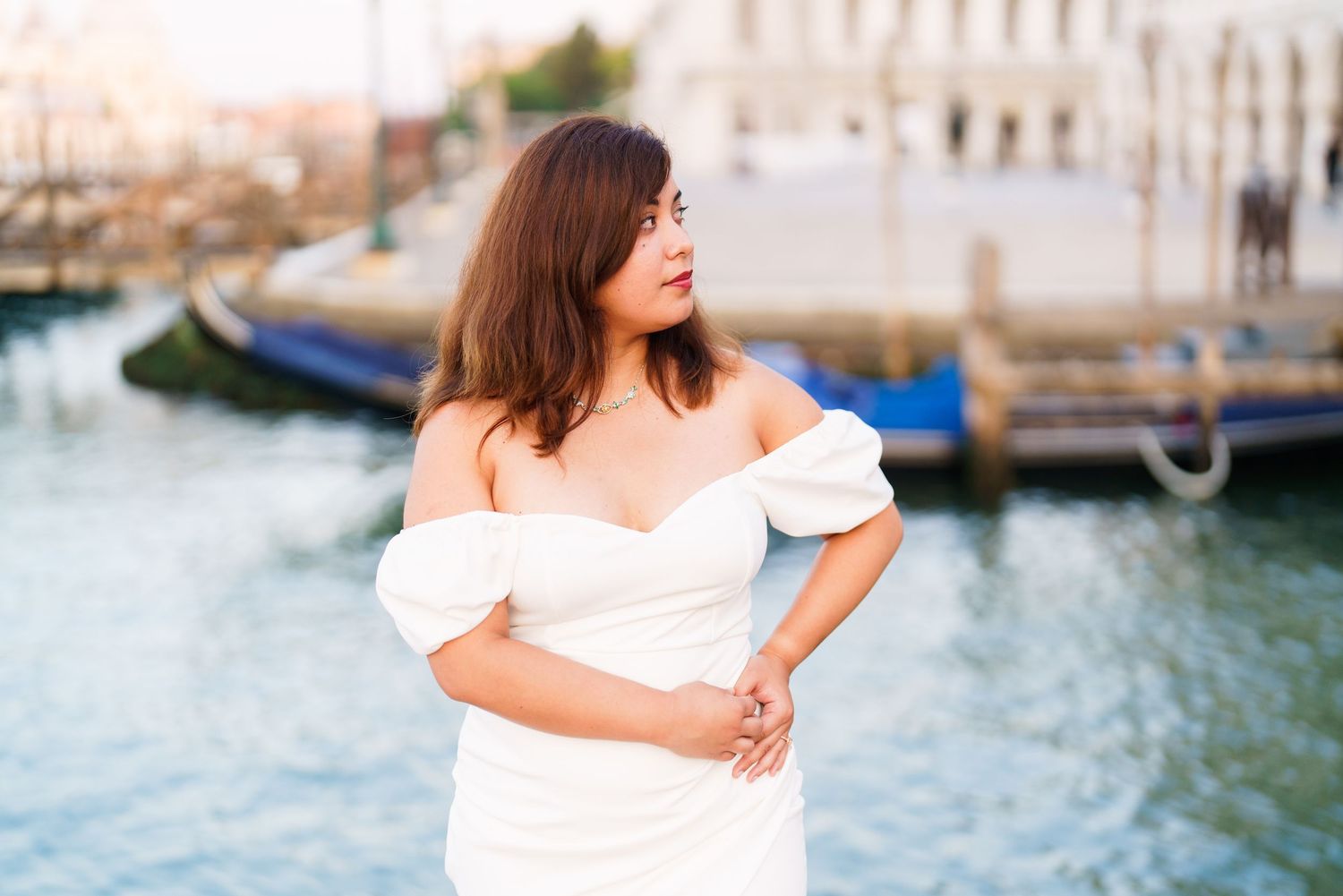 Woman in white dress poses by the Venice canals with boats and historic buildings in the background.
