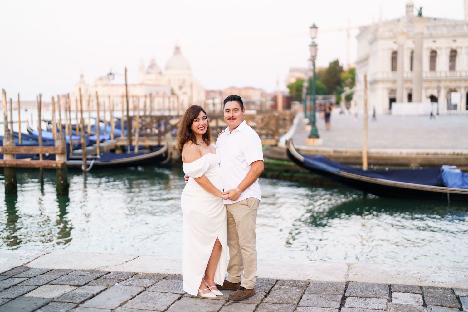 A romantic couple poses for photos along the canals of Venice with gondolas and historic architecture in the background.