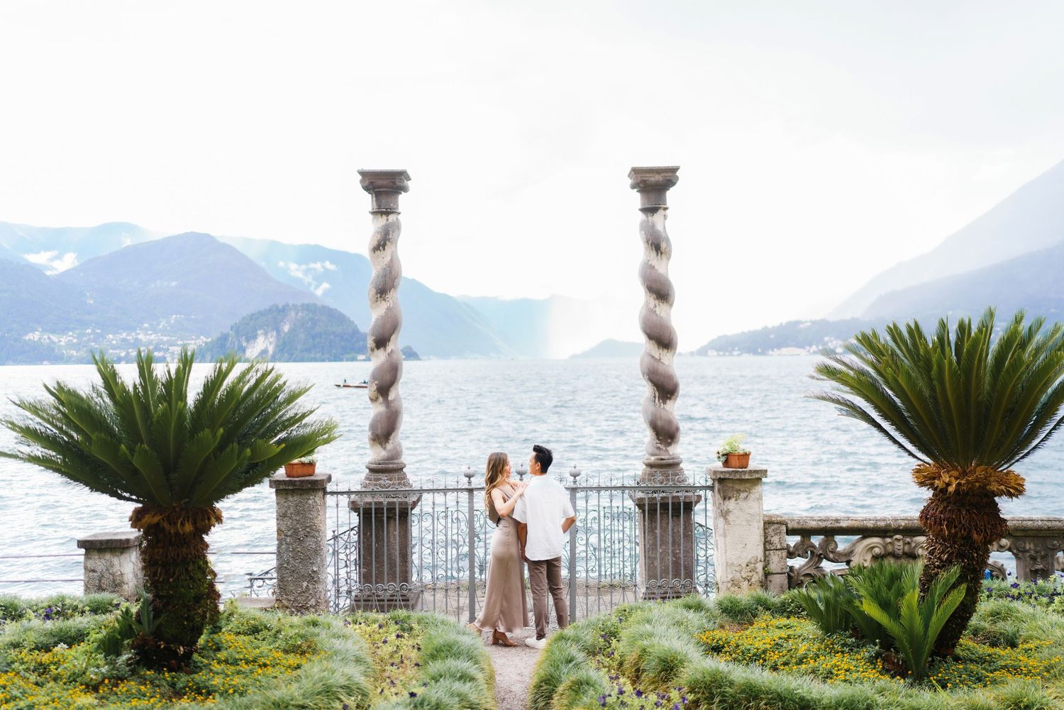 Romantic couple stands between ornate columns overlooking Lake Como in Italy with palm trees and mountains in view.
