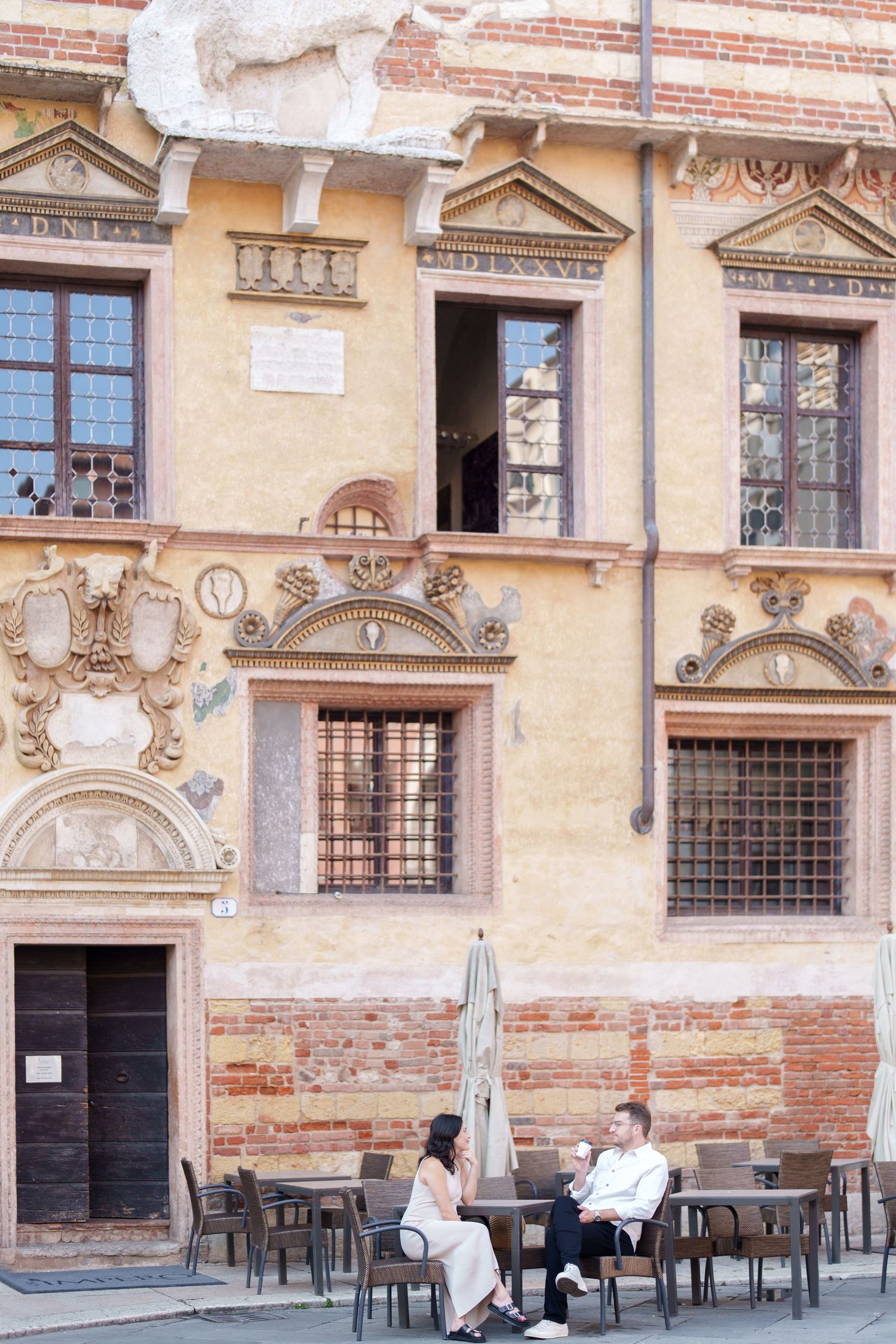Historic Italian cafe terrace with outdoor seating against ornate classical building facade with decorative windows and arches.