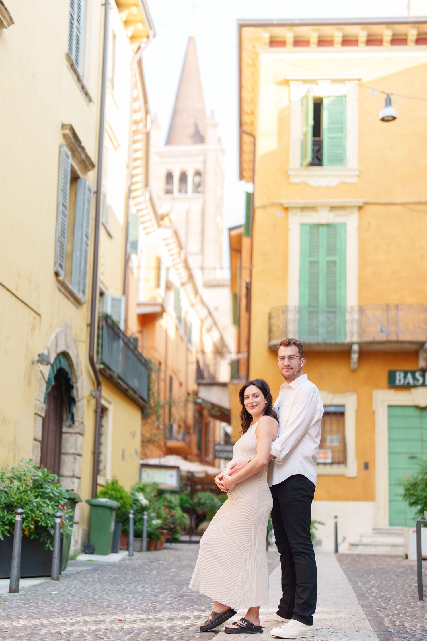 A couple stands together on a charming cobblestone street in Italy with colorful buildings and a church spire in the background.