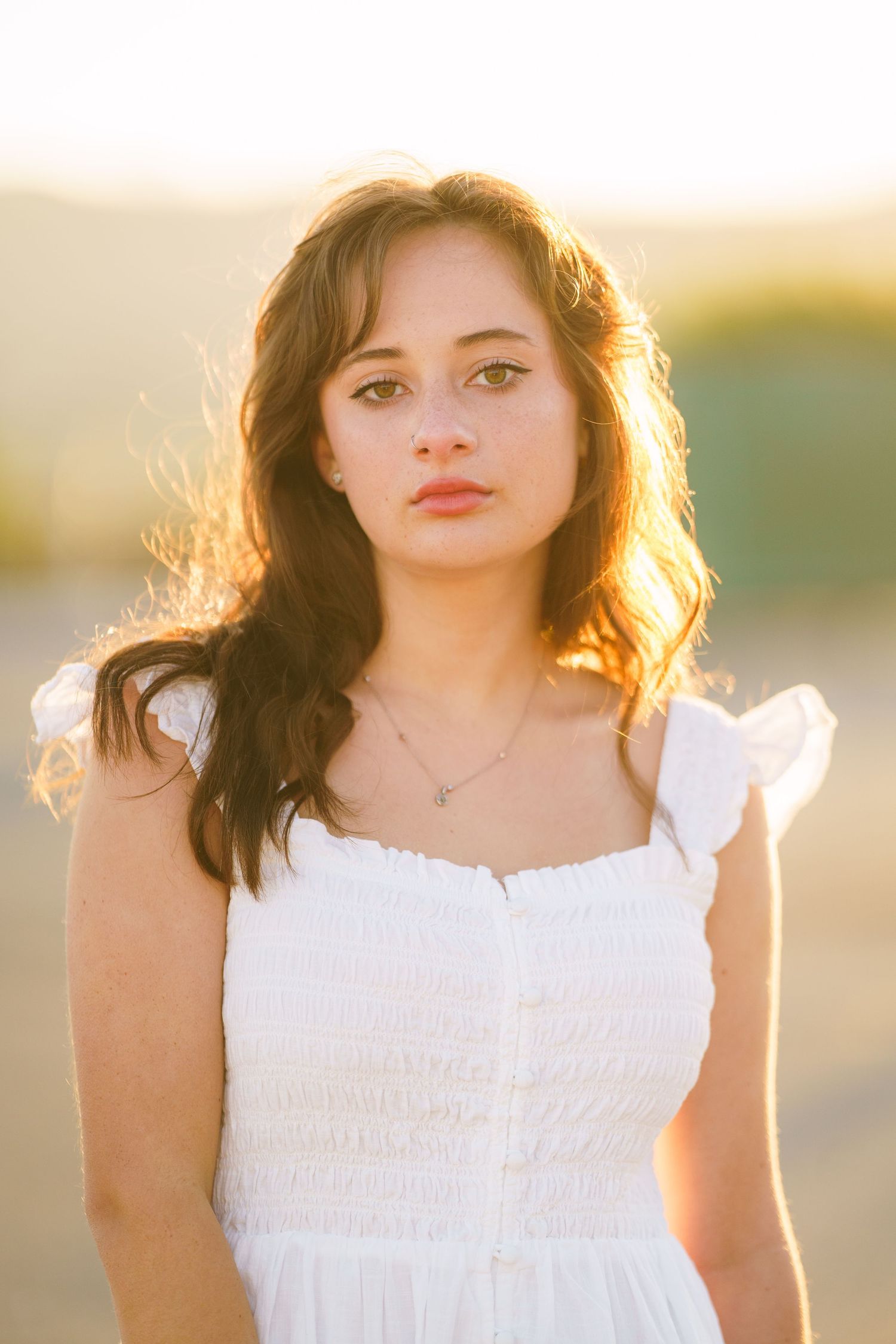 A young woman in a white sundress poses outdoors during golden hour with sunlit hair and natural lighting.