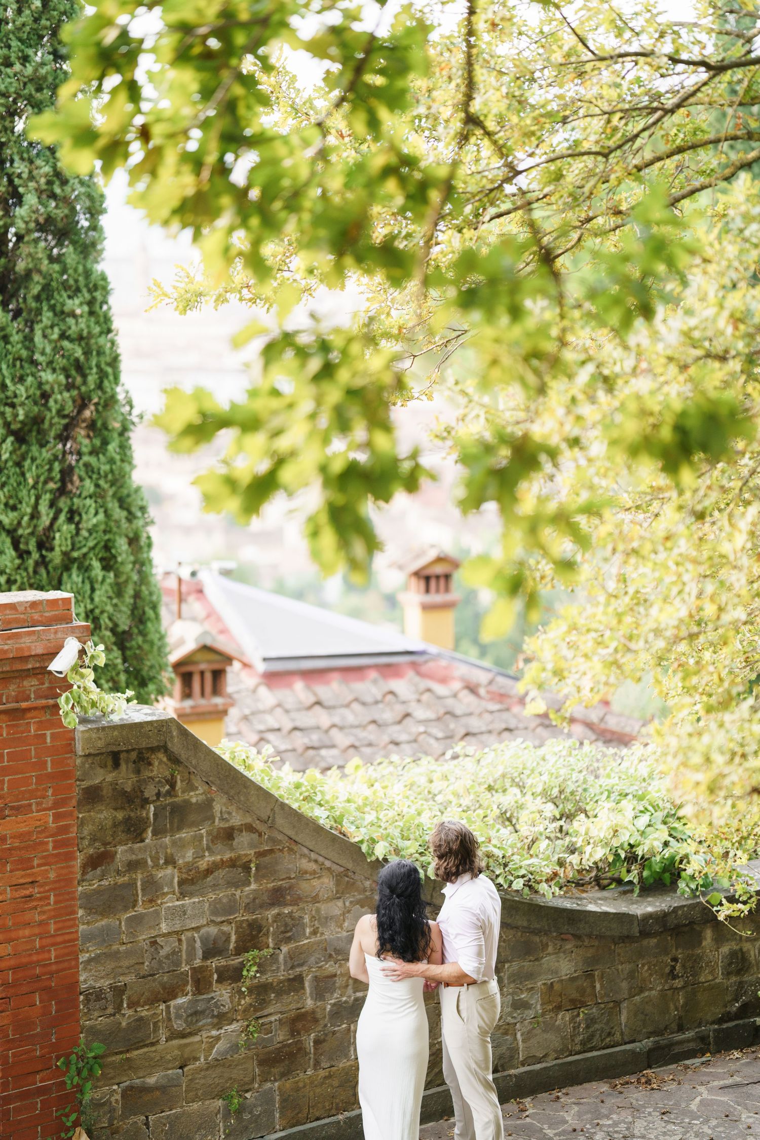 A couple embraces while overlooking red-tiled rooftops through lush green foliage in a romantic garden setting.