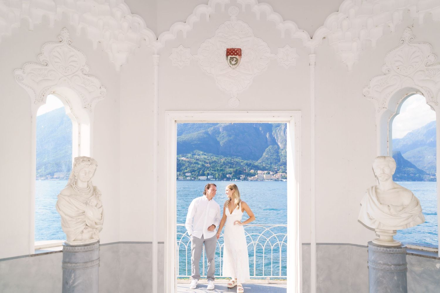 A scenic white balcony overlooking a serene mountain lake with multiple architectural columns framing the view.