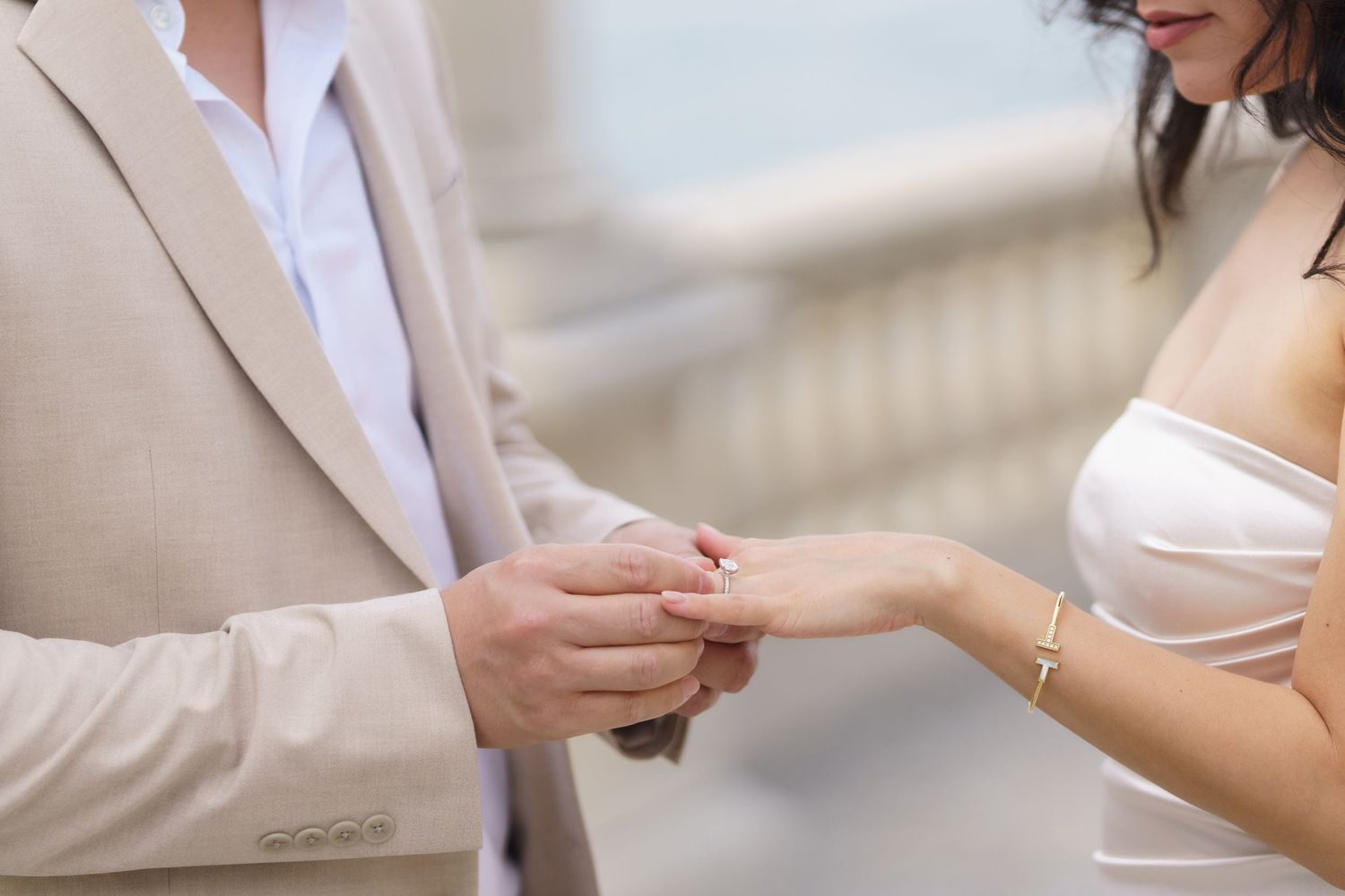 Close-up of hands during wedding ceremony as groom places ring on bride's finger wearing beige suit and white dress.