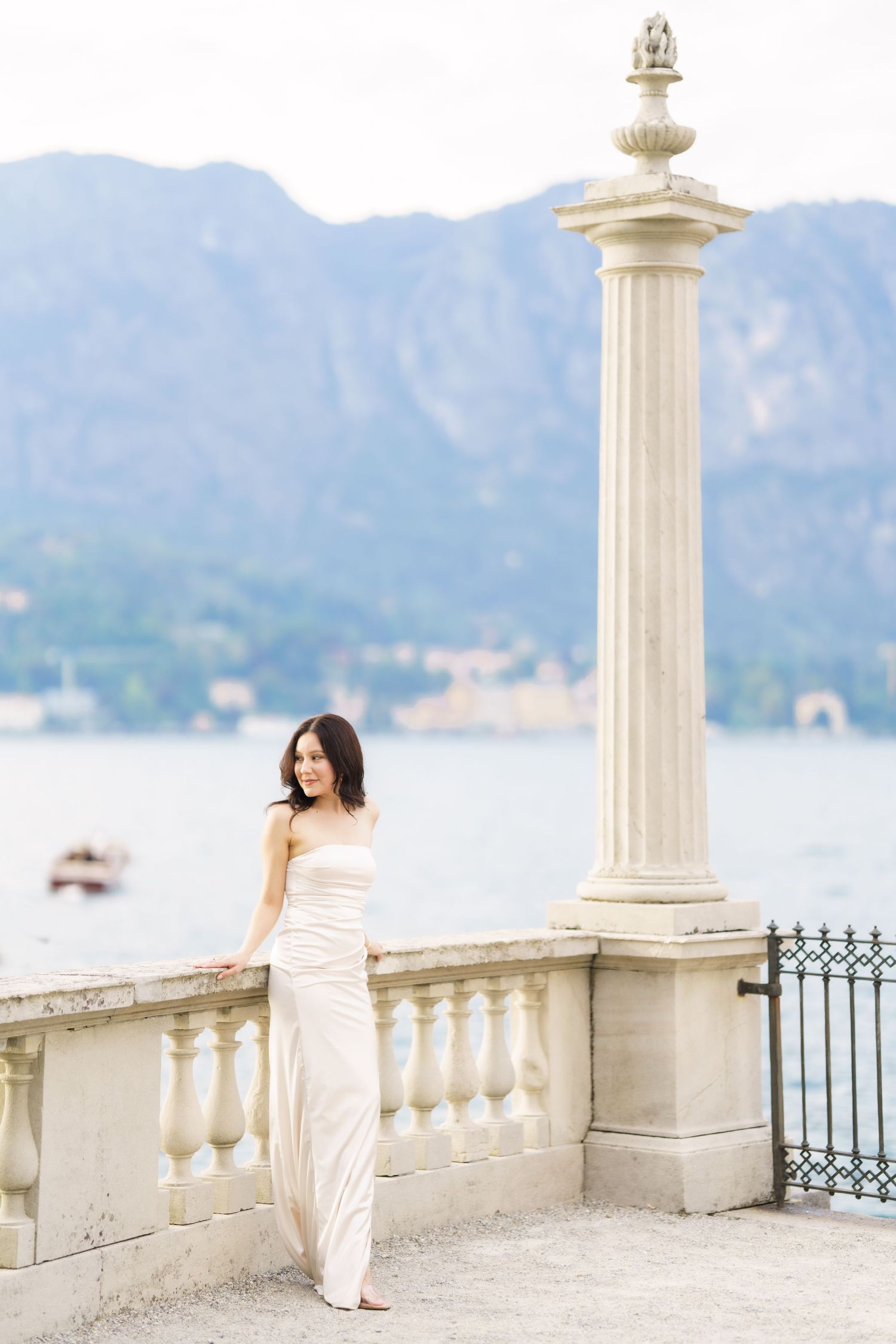 A person in a white dress stands on an elegant stone terrace overlooking a scenic lake and mountains.
