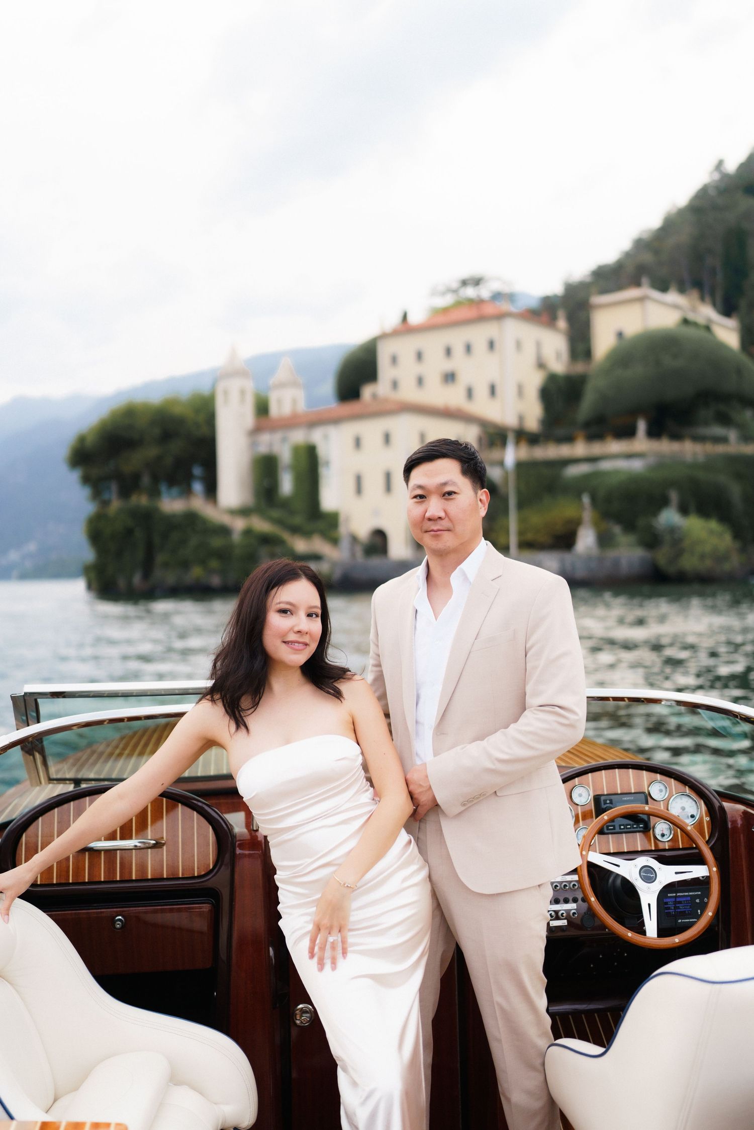 Romantic couple poses in elegant white attire on a vintage wooden boat near a Mediterranean coastal villa.