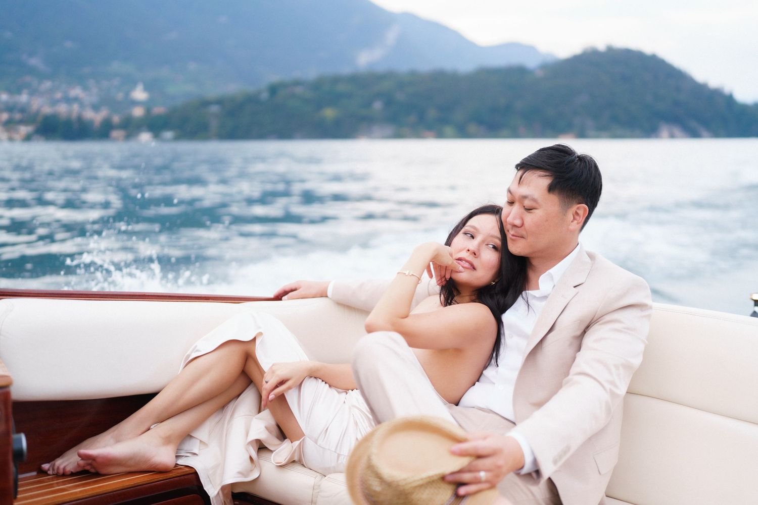 A couple in white attire relaxes together on a boat with mountains and lake scenery in the background.