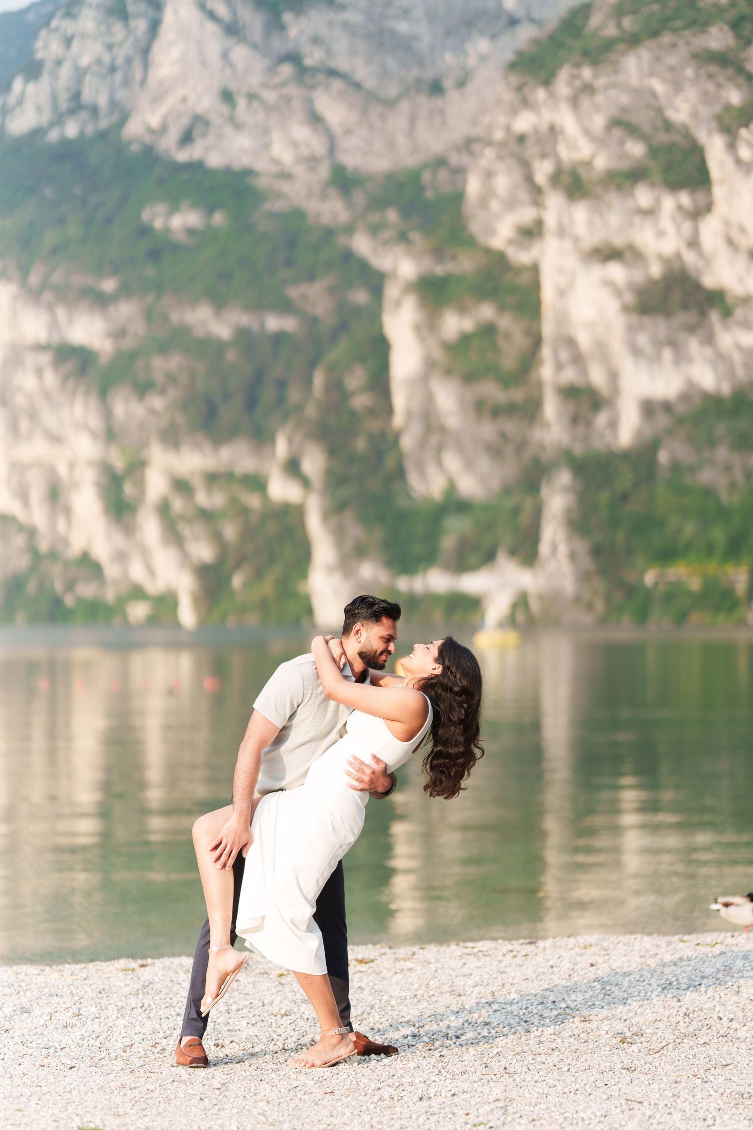 A romantic couple shares an intimate moment on a pebble beach with dramatic mountain cliffs and a serene lake in the background.