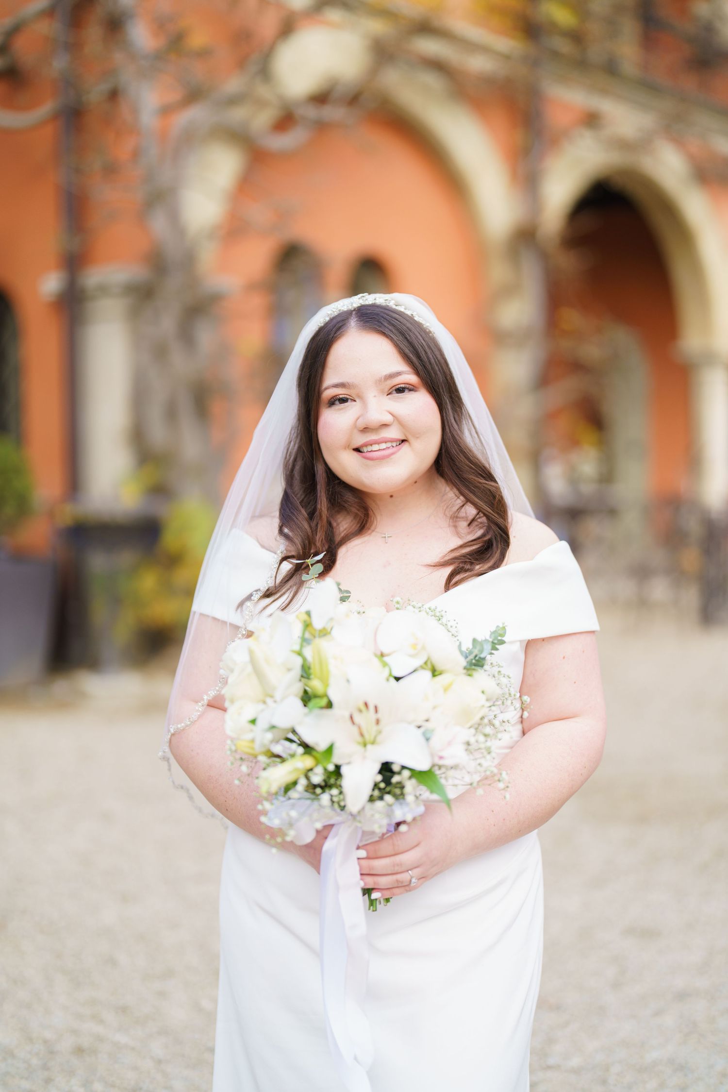 A bride in a white dress smiles while holding a bouquet of white flowers against an architectural backdrop.