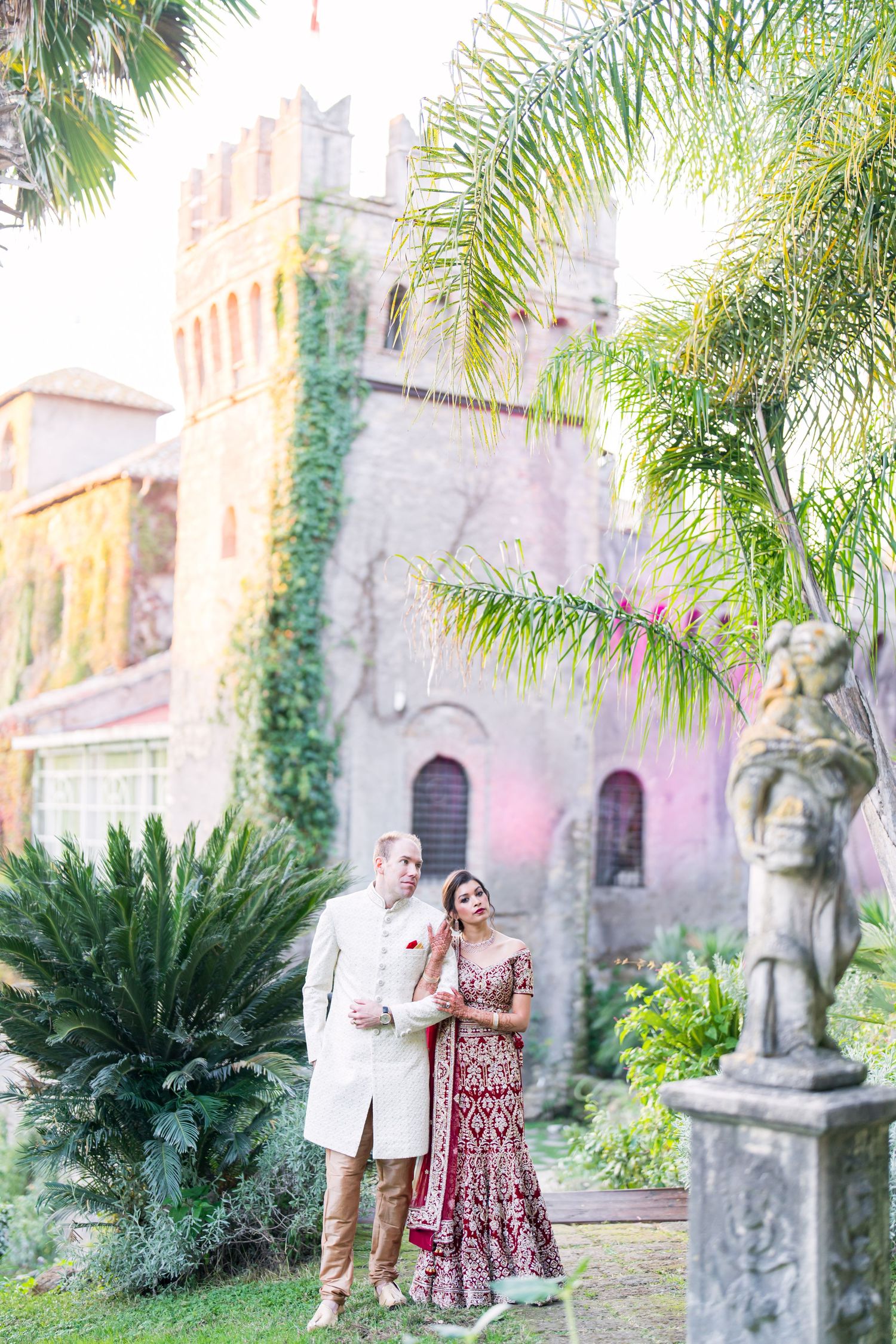 A couple shares an intimate moment in a garden with palm trees and stone architecture in the background.