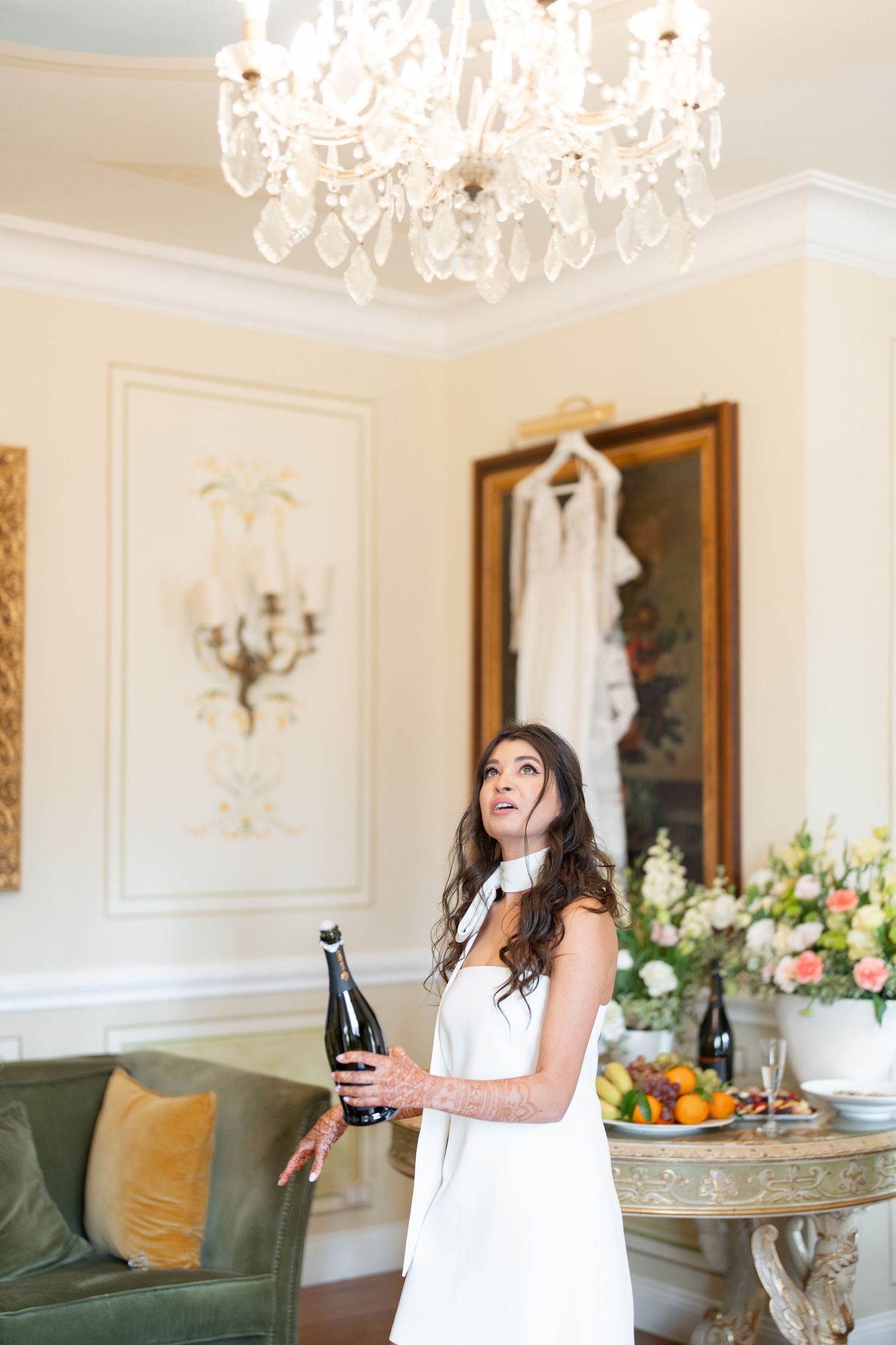 Woman in white dress celebrates with champagne in elegant room with chandelier and floral decor.