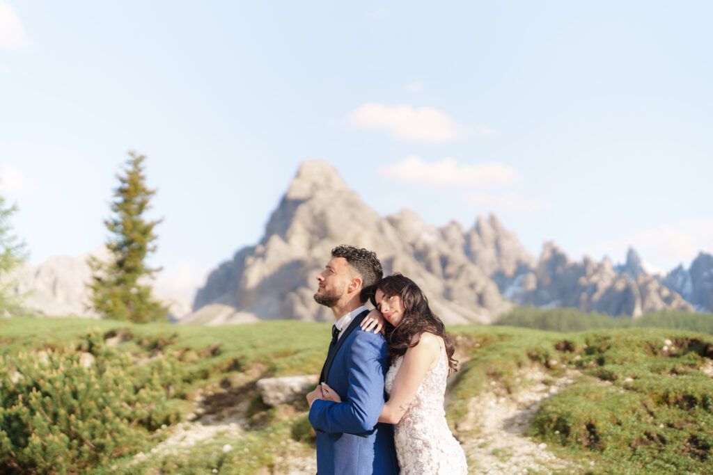 Two people embrace on a scenic mountain overlook with majestic alpine peaks behind them.