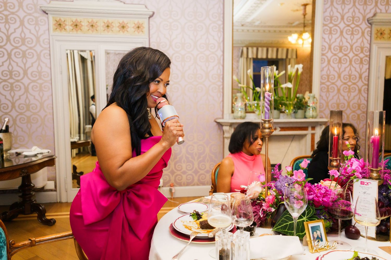 Woman in pink dress giving a toast at an elegant dinner party with floral centerpieces and candlelight.