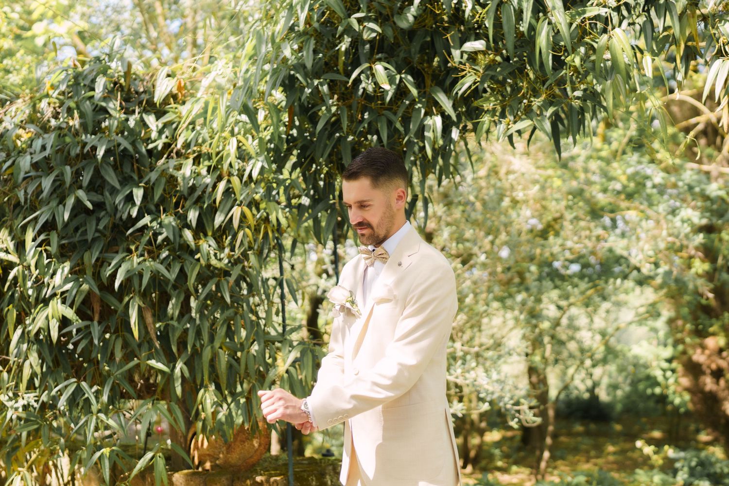 Groom in a cream suit adjusting his cufflinks outdoors among lush green trees on his wedding day.