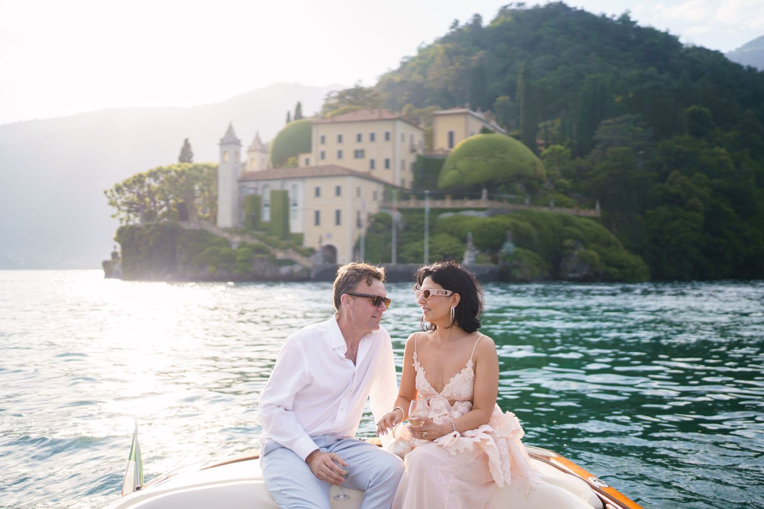 Couple sitting on a boat at Lake Como with Villa del Balbianello in the background on a sunny day.