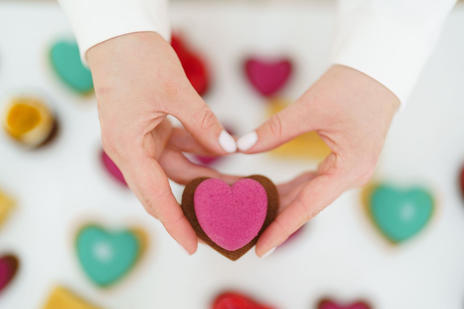 Hands holding a pink heart-shaped chocolate cookie with colorful heart cookies in the background.