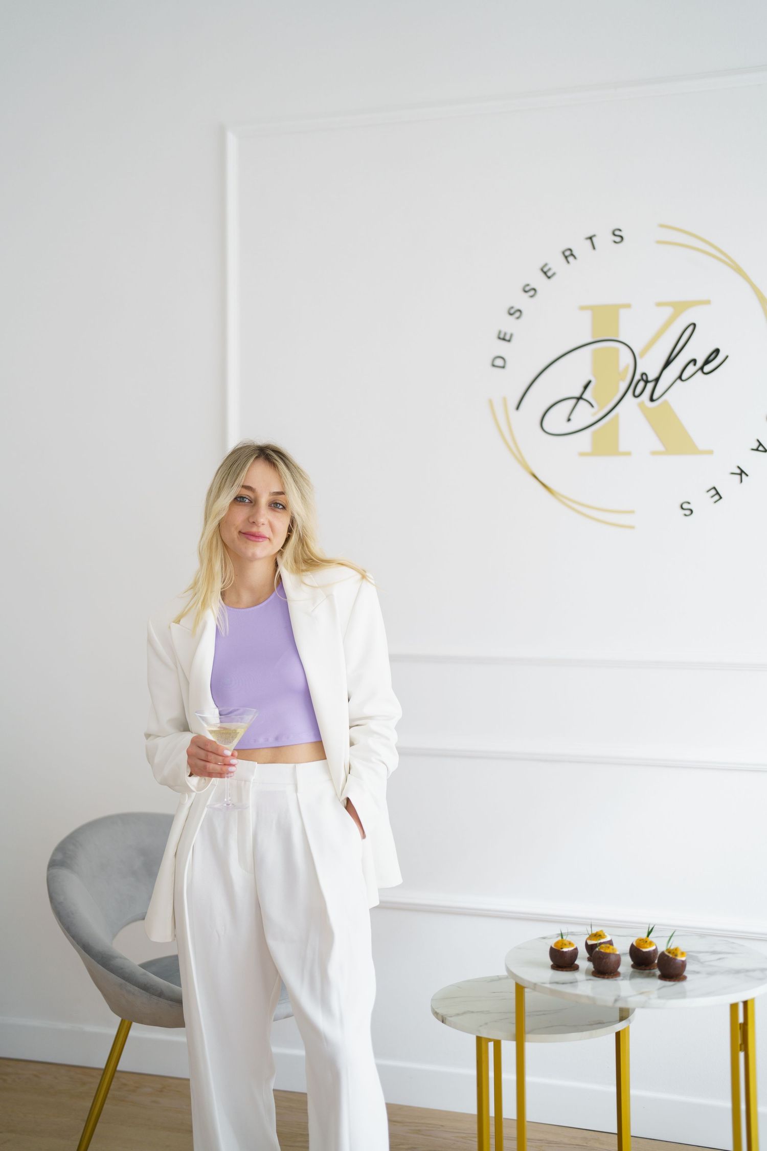 Woman in white suit holding a glass stands near Dolce K Desserts Cakes logo sign with cupcakes on gold table.