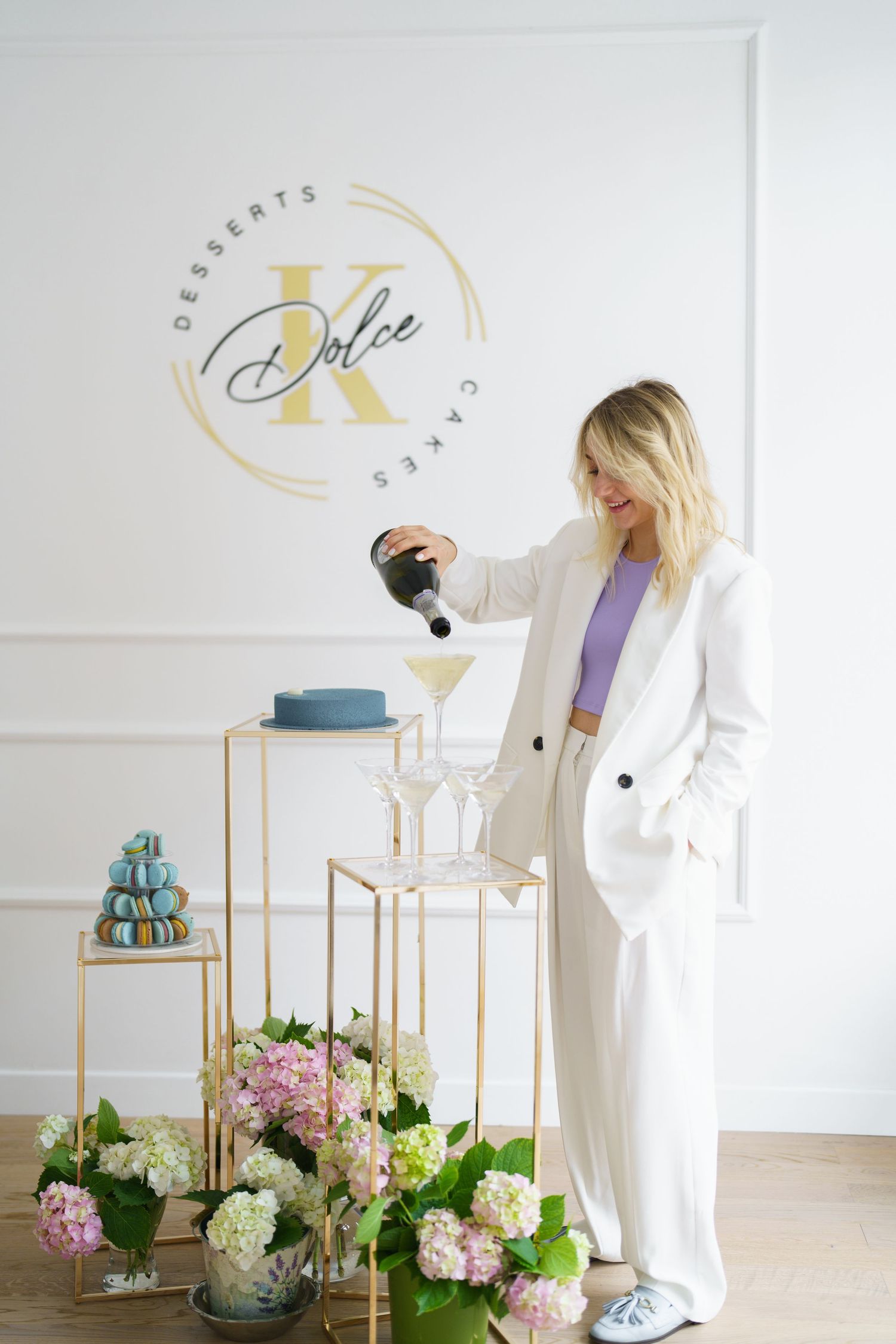Woman in white suit pouring champagne at Dolce K Desserts & Cakes event with floral arrangements and macarons.