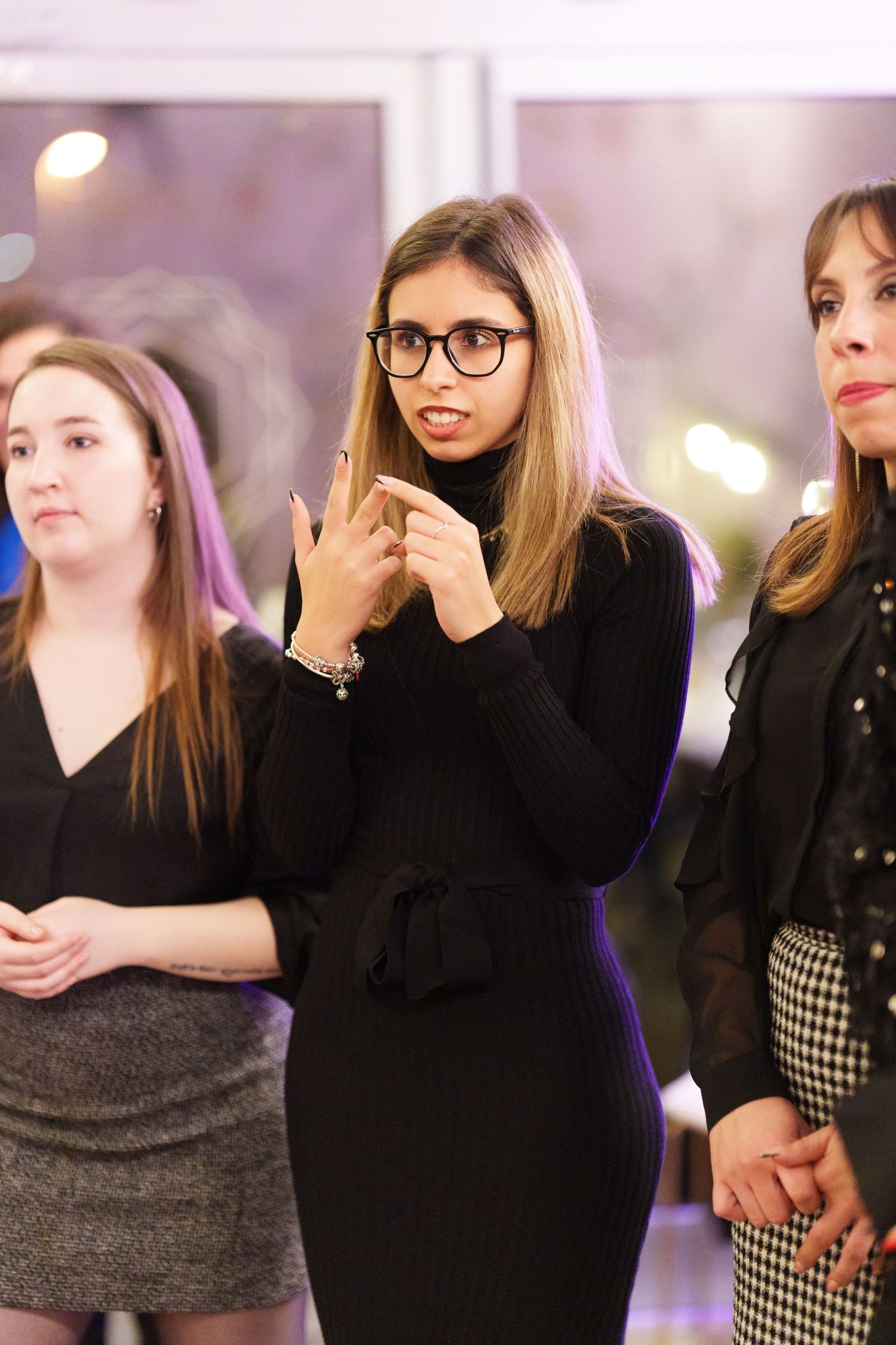 Three women in black outfits standing together at an indoor event, center woman wearing glasses with hands gesturing.