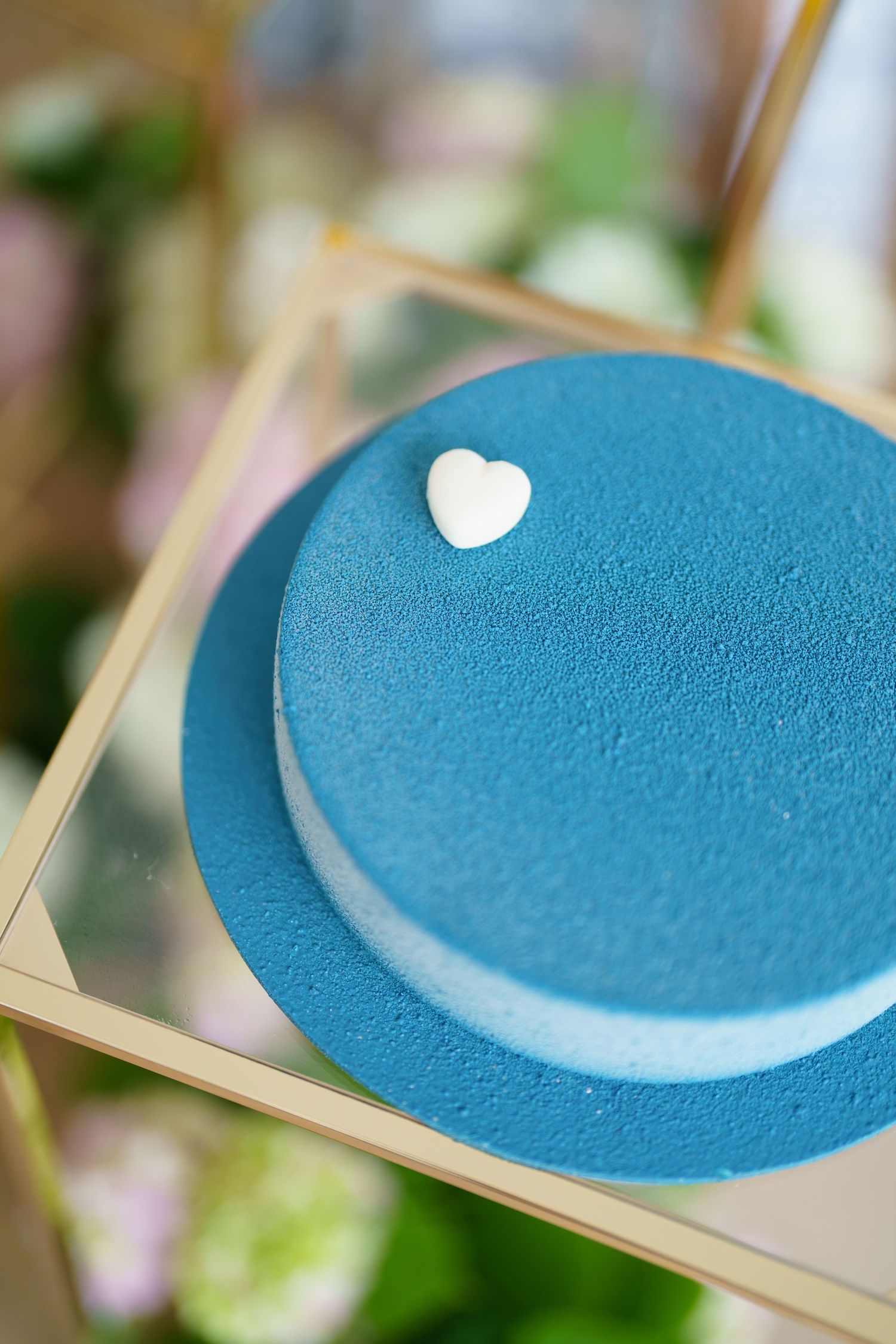 Blue velvet cake with white heart decoration on gold tray, surrounded by flowers.