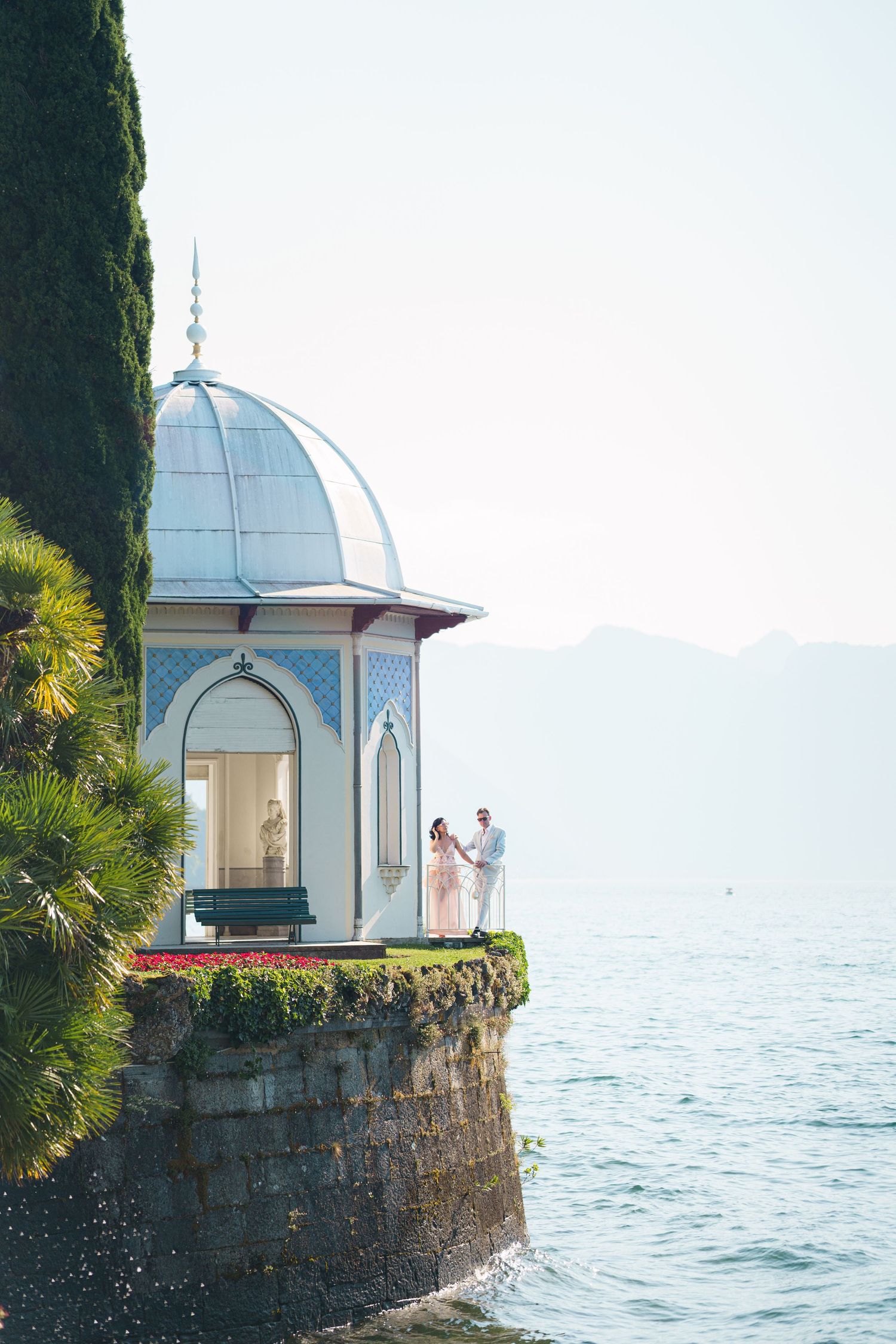 Couple standing on ornate lakeside pavilion with blue dome, surrounded by lush greenery and calm water with mountains beyond.